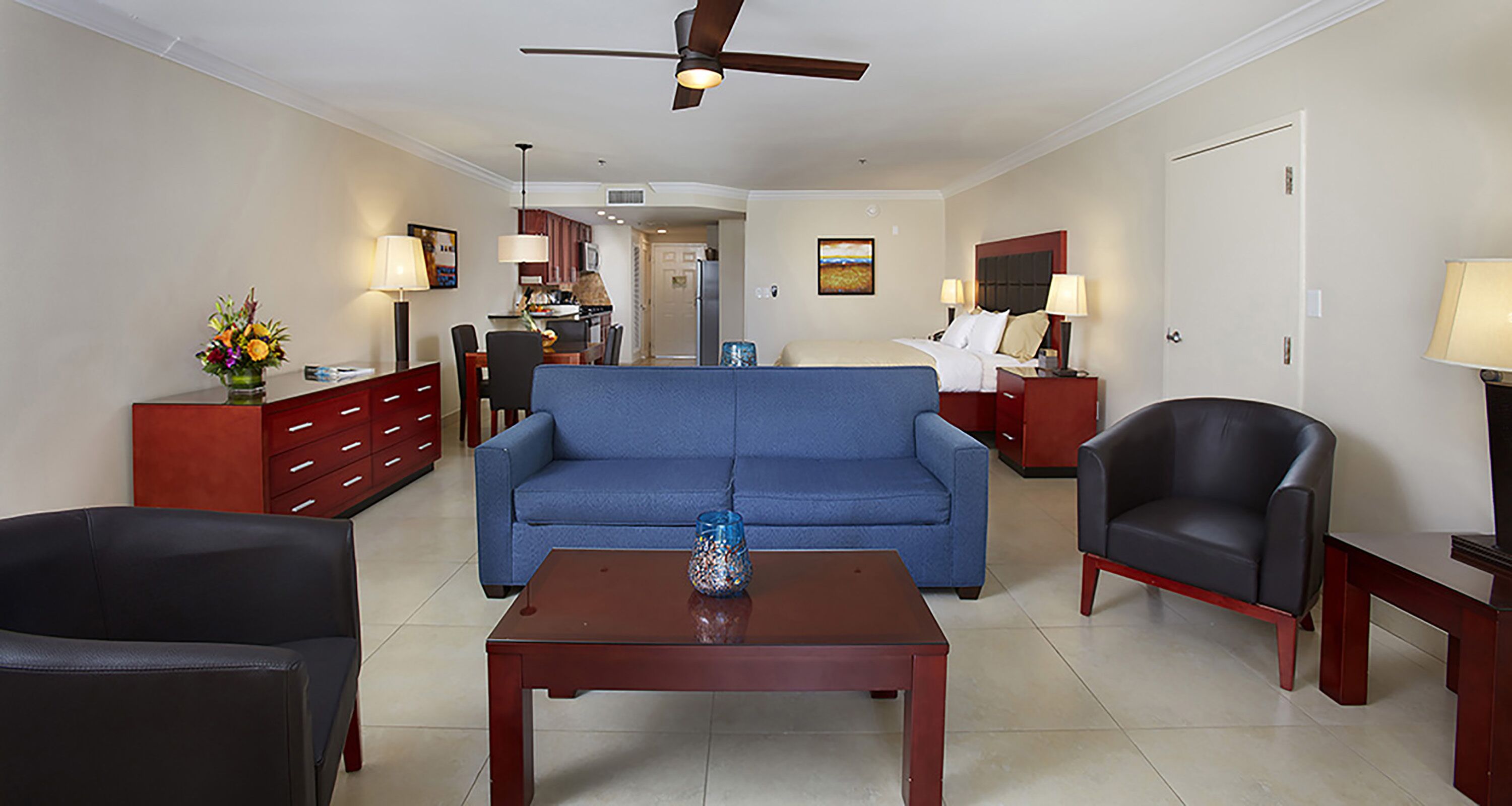 A hotel room with a blue sofa, a black armchair, a wooden coffee table, a red side table, a TV, and a ceiling fan.