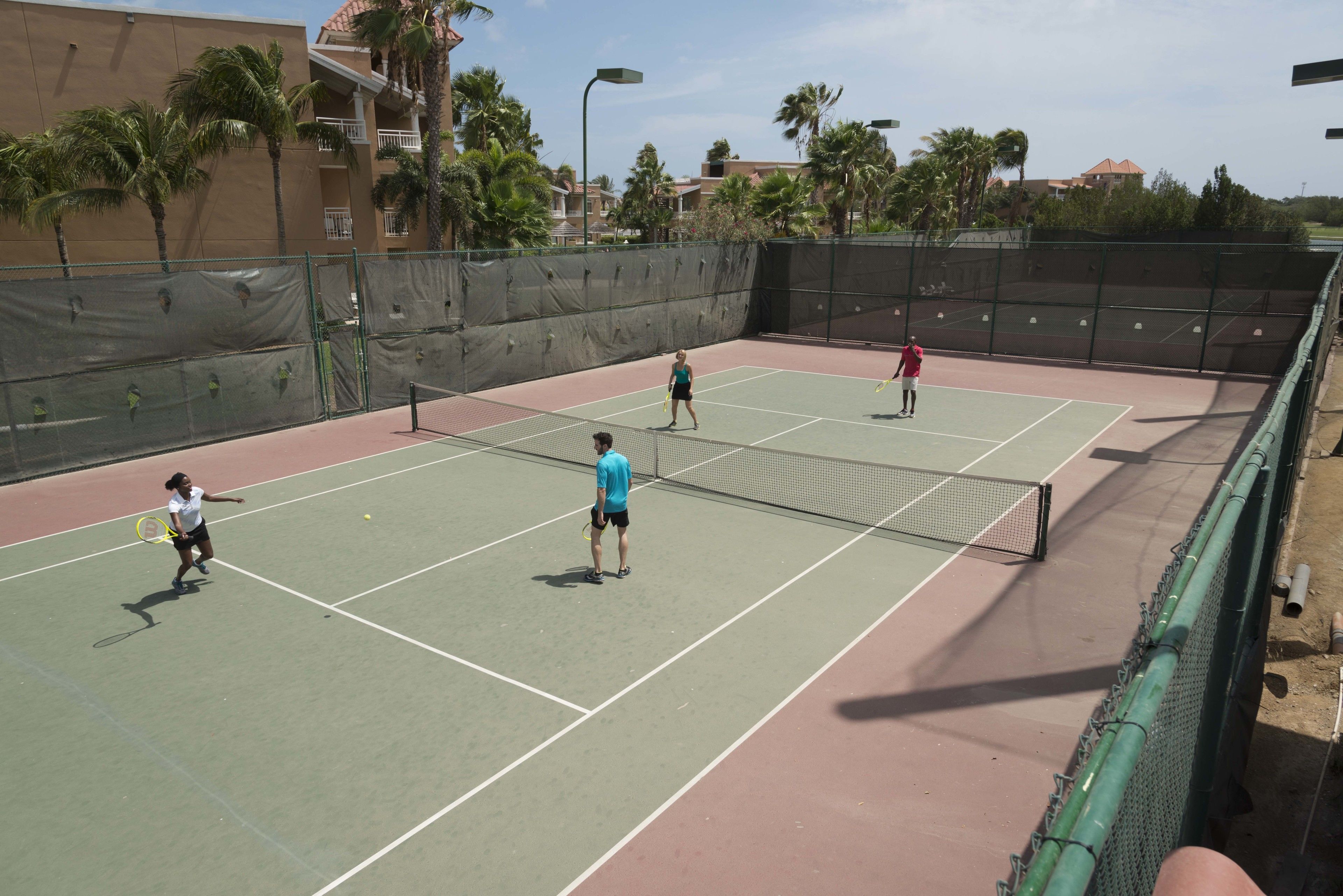 A tennis court with a net, surrounded by a fence and palm trees.