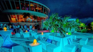 An outdoor seating area with white cushioned chairs and tables, surrounded by palm trees and a building with glass windows in the background.