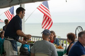 Un espace repas avec des tables et des chaises, des drapeaux américains et une vue sur l’océan.