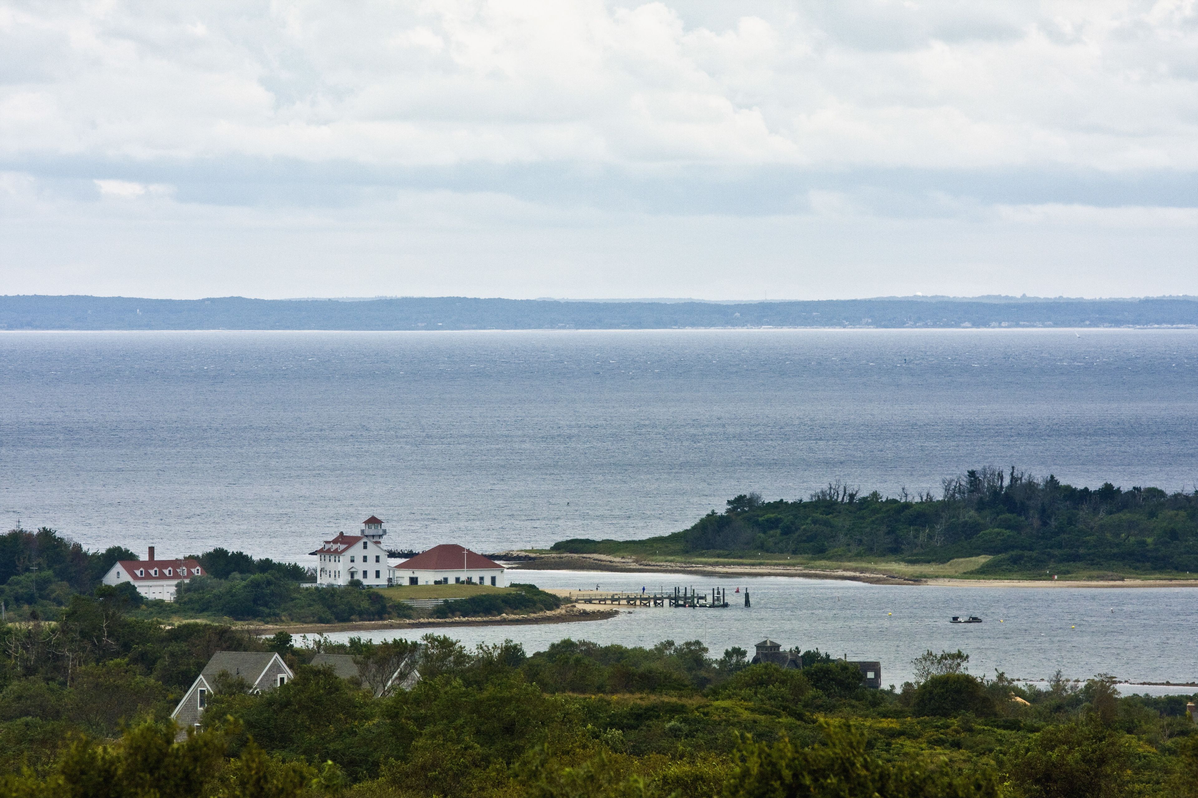 A coastal view with a white building with a red roof, a pier extending into the water, and a distant shoreline.