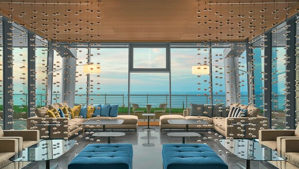 A modern lounge with sofas and tables, large windows, and hanging lights.