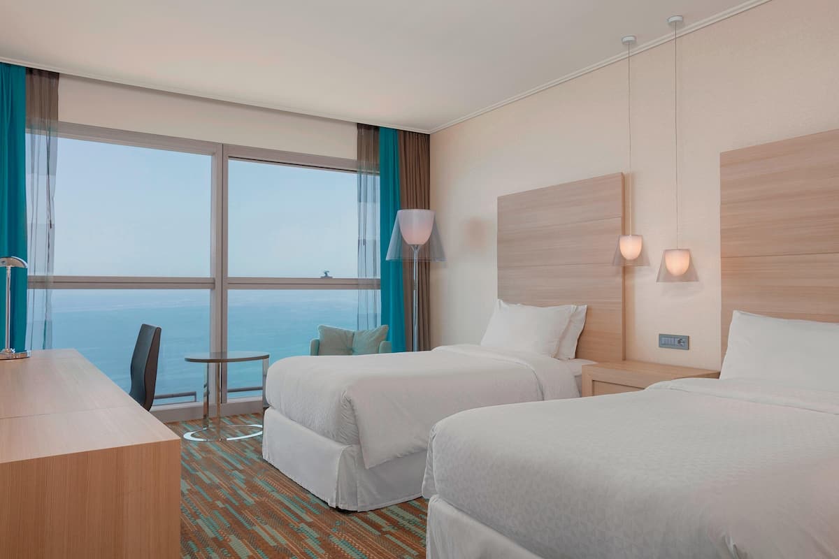 A hotel room with two beds, a desk, a small table, and a view of the ocean.