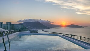 A rooftop infinity pool with a view of the sunset over the sea and mountains.