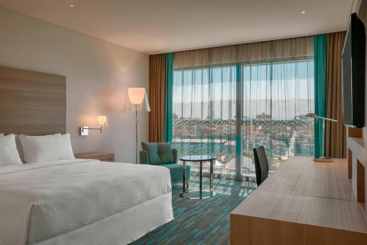 A hotel room with a large bed, a desk, a chair, a small table, a lamp, and a view of the city through large windows.
