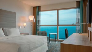 A hotel room with a large bed, a desk, a chair, a small table, and a TV. The room has a view of the ocean.