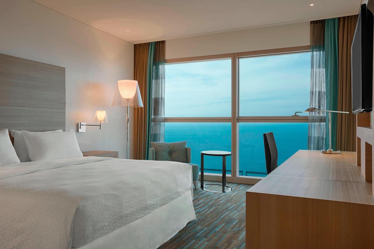 A hotel room with a large bed, a desk, a chair, a small table, and a TV. The room has a view of the ocean.