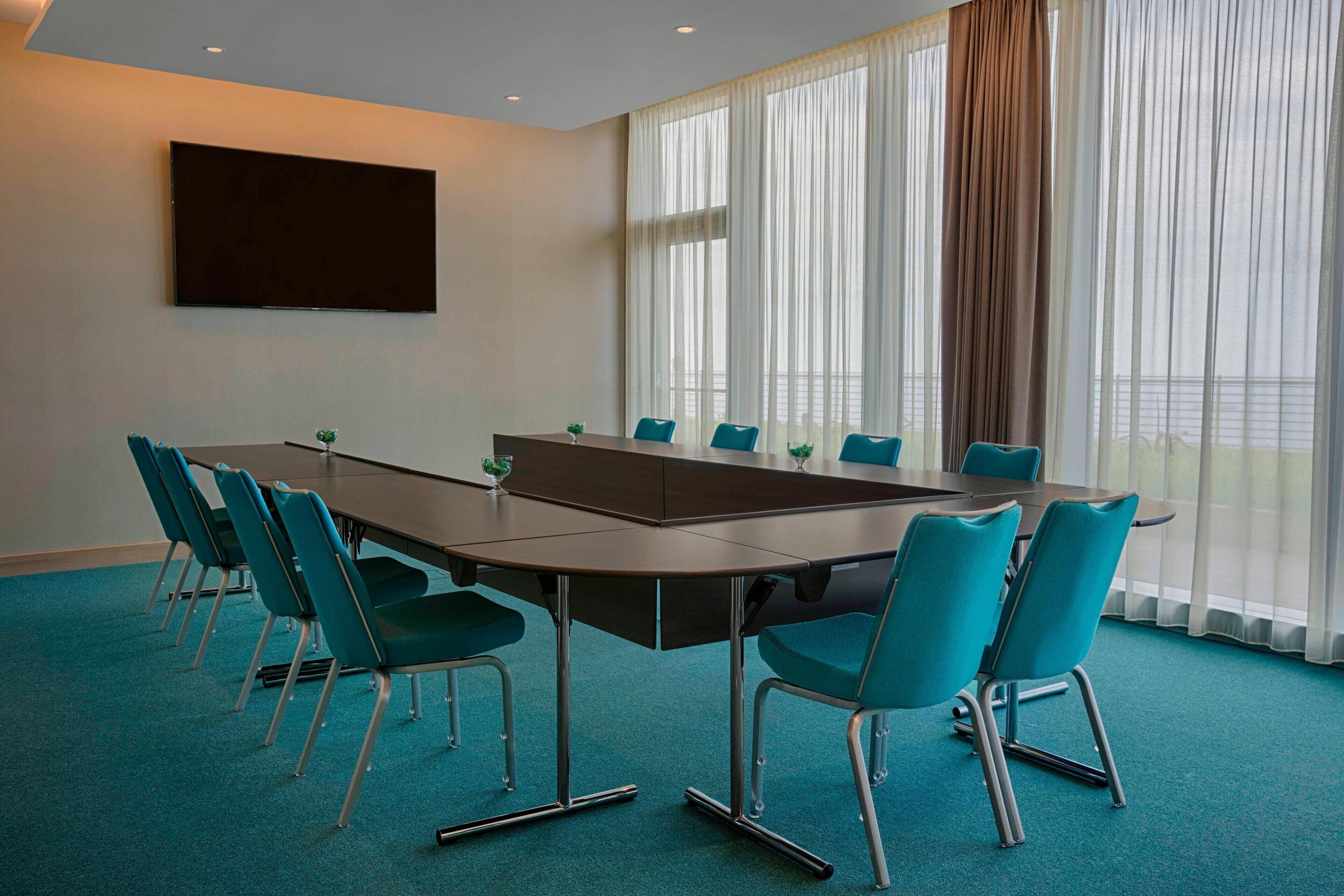 A conference room with a long table, teal chairs, a flat-screen TV, and large windows with sheer curtains.