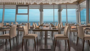 A restaurant with tables and chairs arranged for dining, large windows offering a view of the sea, and a ceiling with a unique, scalloped design.