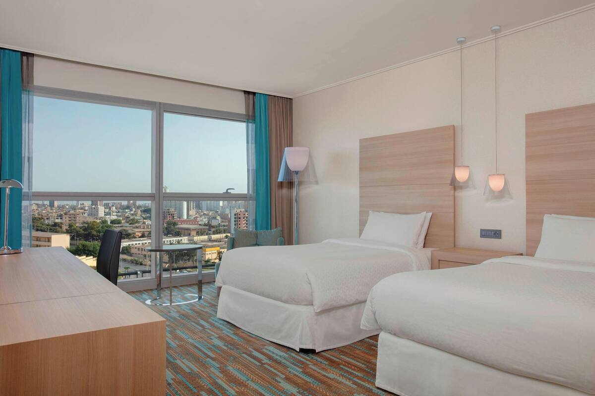 A hotel room with two beds, a desk, a chair, a lamp, and a view of the city.