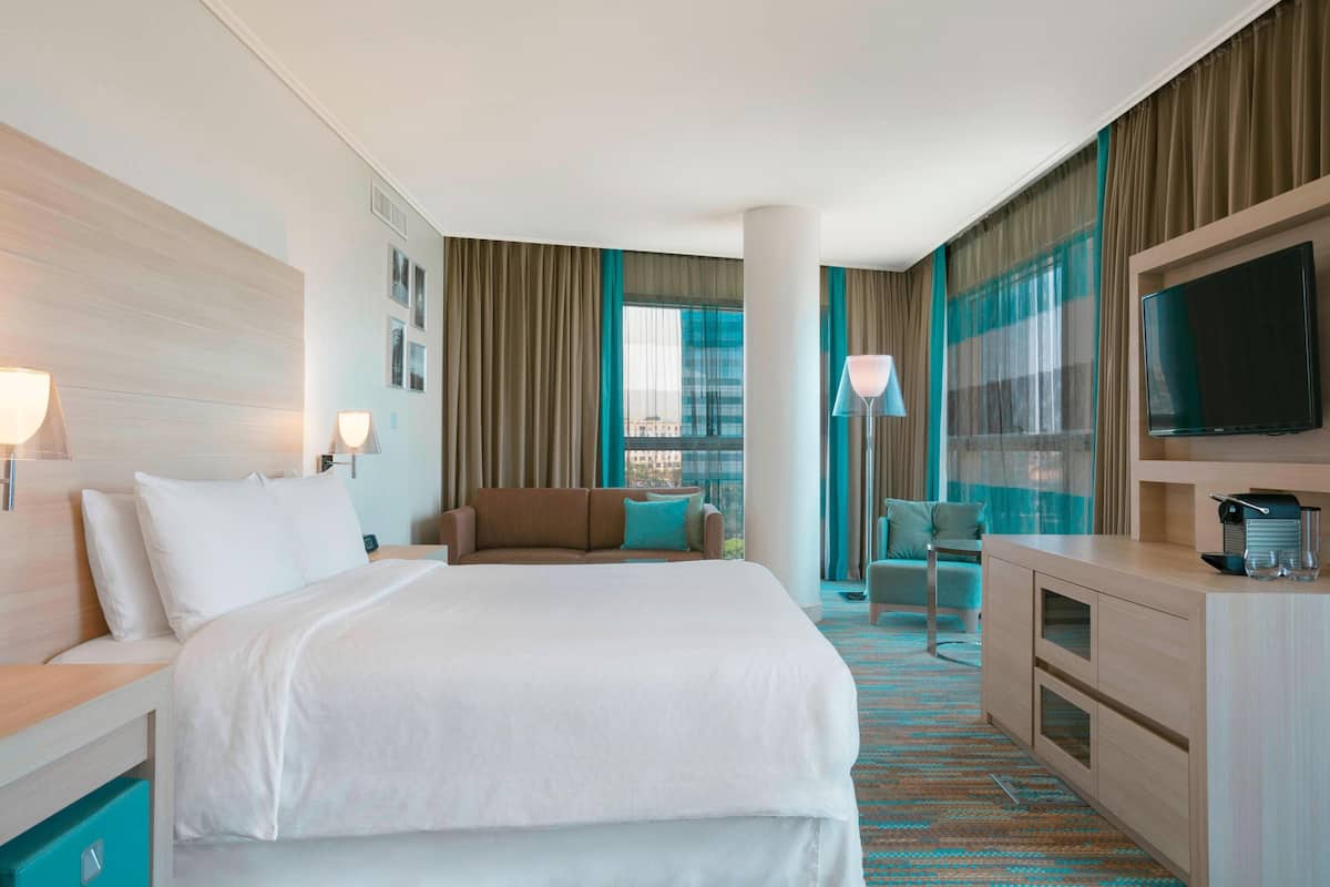 A hotel room with a large bed, a sofa, a desk with a TV, and a view of the city through large windows.