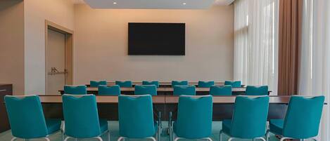 Ein Konferenzraum mit türkisfarbenen Stühlen, einem langen Tisch und einer großen Leinwand an der Wand.