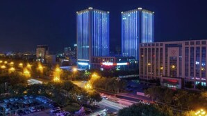 A nighttime cityscape with two illuminated skyscrapers, a parking lot, and a road with moving vehicles. - Blue Horizon Hotel (Fuqian Street Dongcheng Dongying) (Dongying)