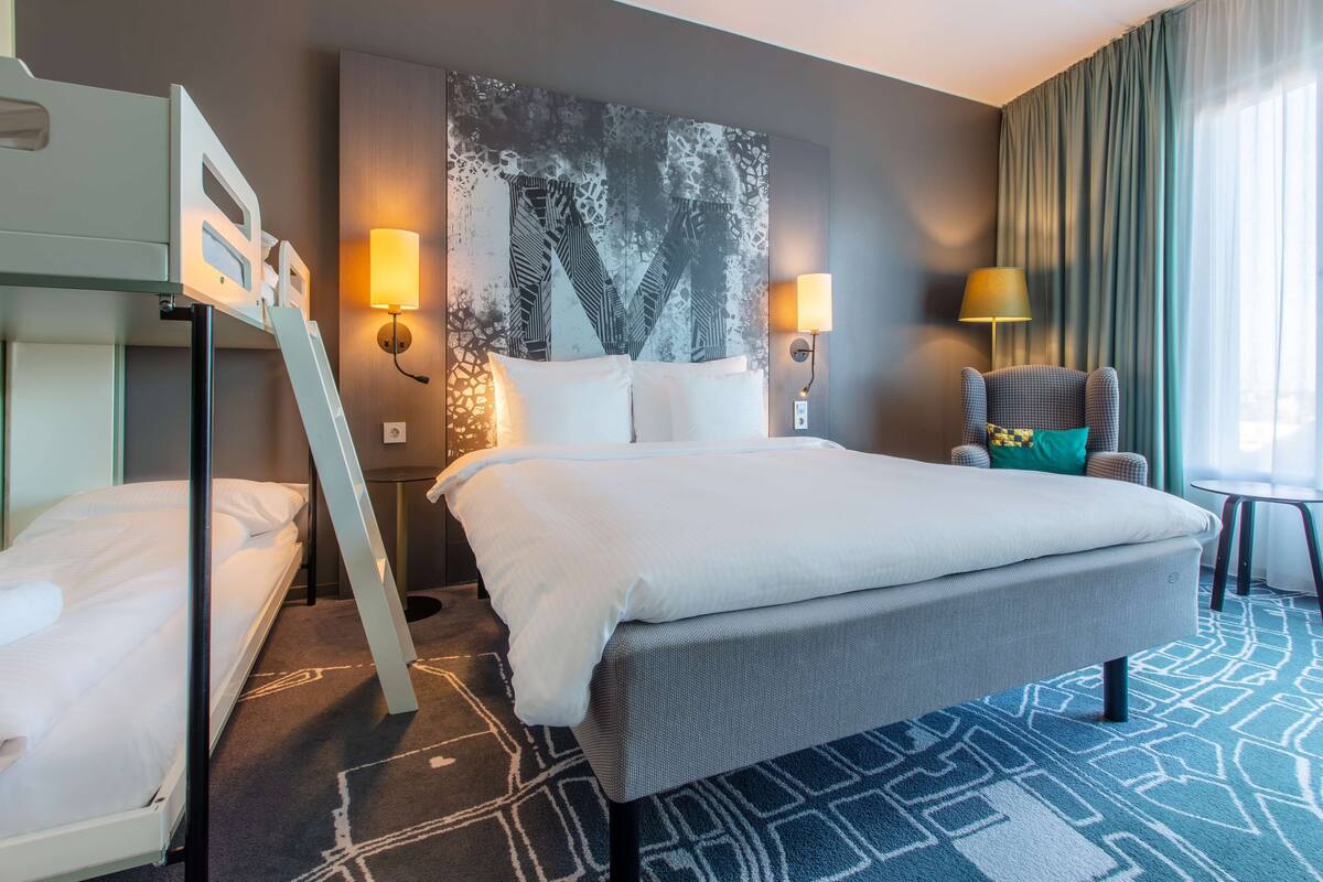A hotel room with a large bed, a bunk bed, two wall-mounted lamps, a chair, and a large abstract painting on the wall.