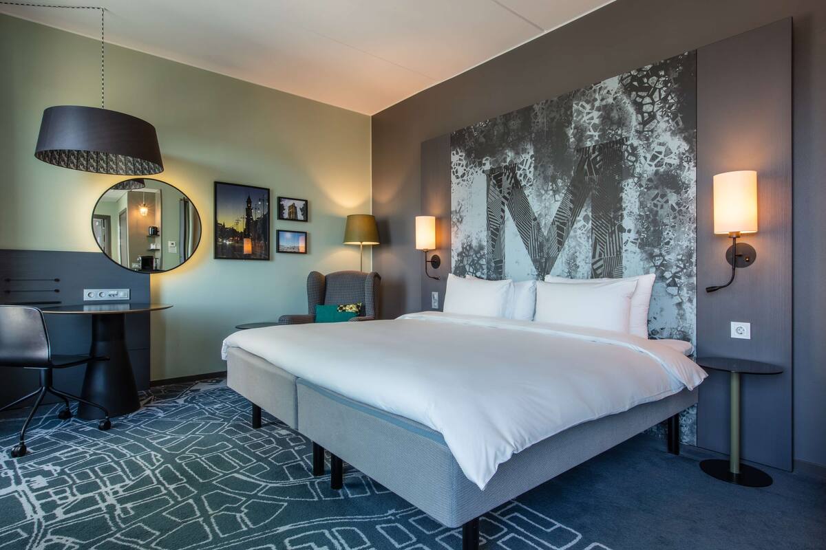 A hotel room with a large bed, a desk, a chair, a round mirror, and two wall-mounted lamps.