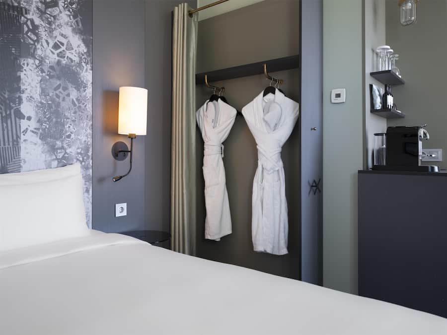 A hotel room with a bed, a wall-mounted light, two white robes hanging on a rack, and a coffee machine on a counter.