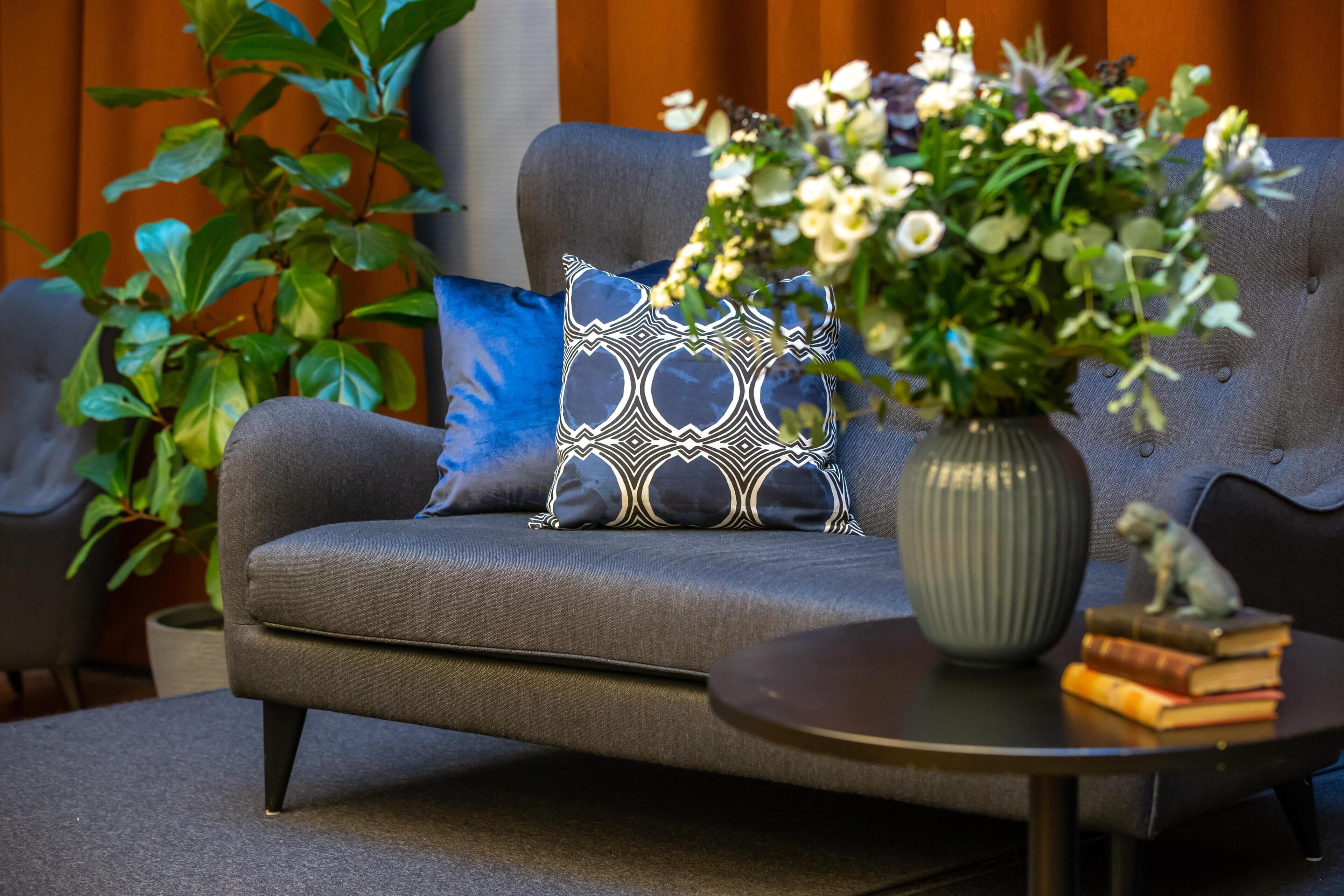 A grey sofa with a patterned pillow, a vase with flowers, and a stack of books on a small table.