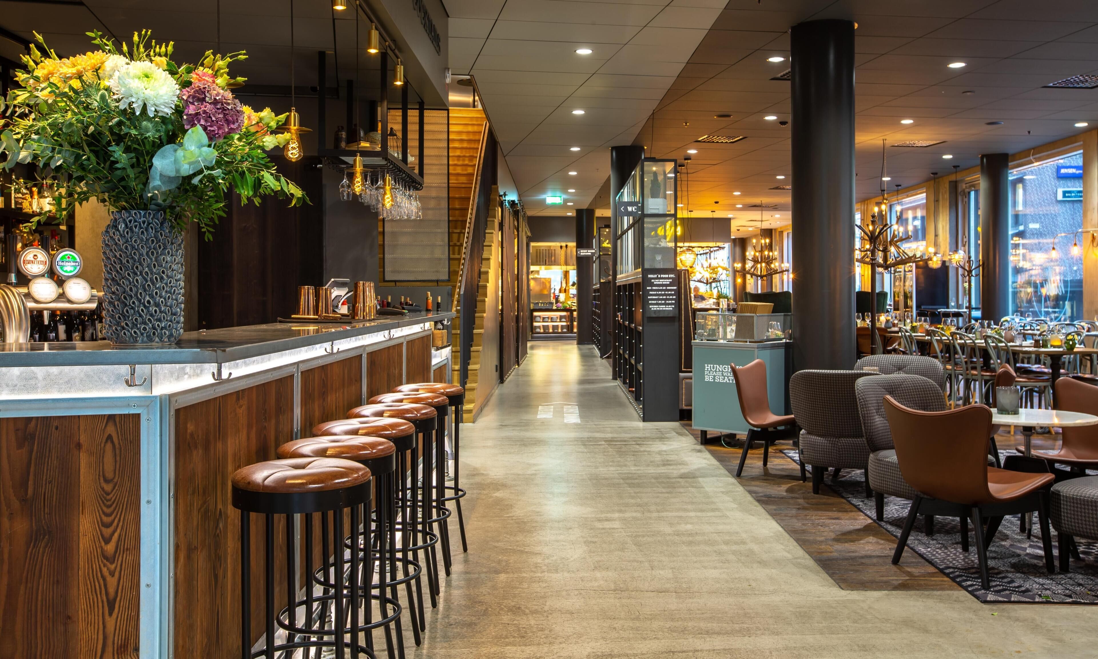 A bar area with stools, a counter with a large vase of flowers, and a view of the dining area with tables and chairs.