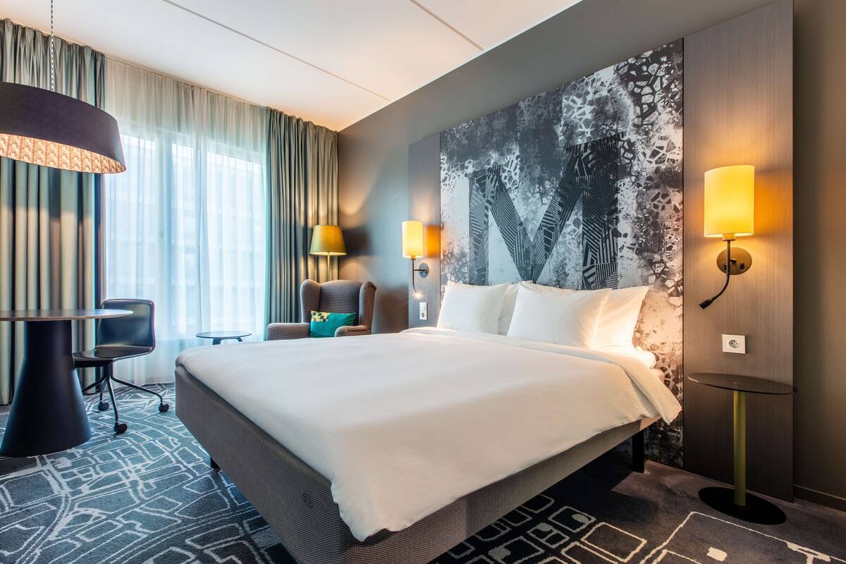 A modern hotel room with a large bed, two bedside lamps, a desk, and a chair.