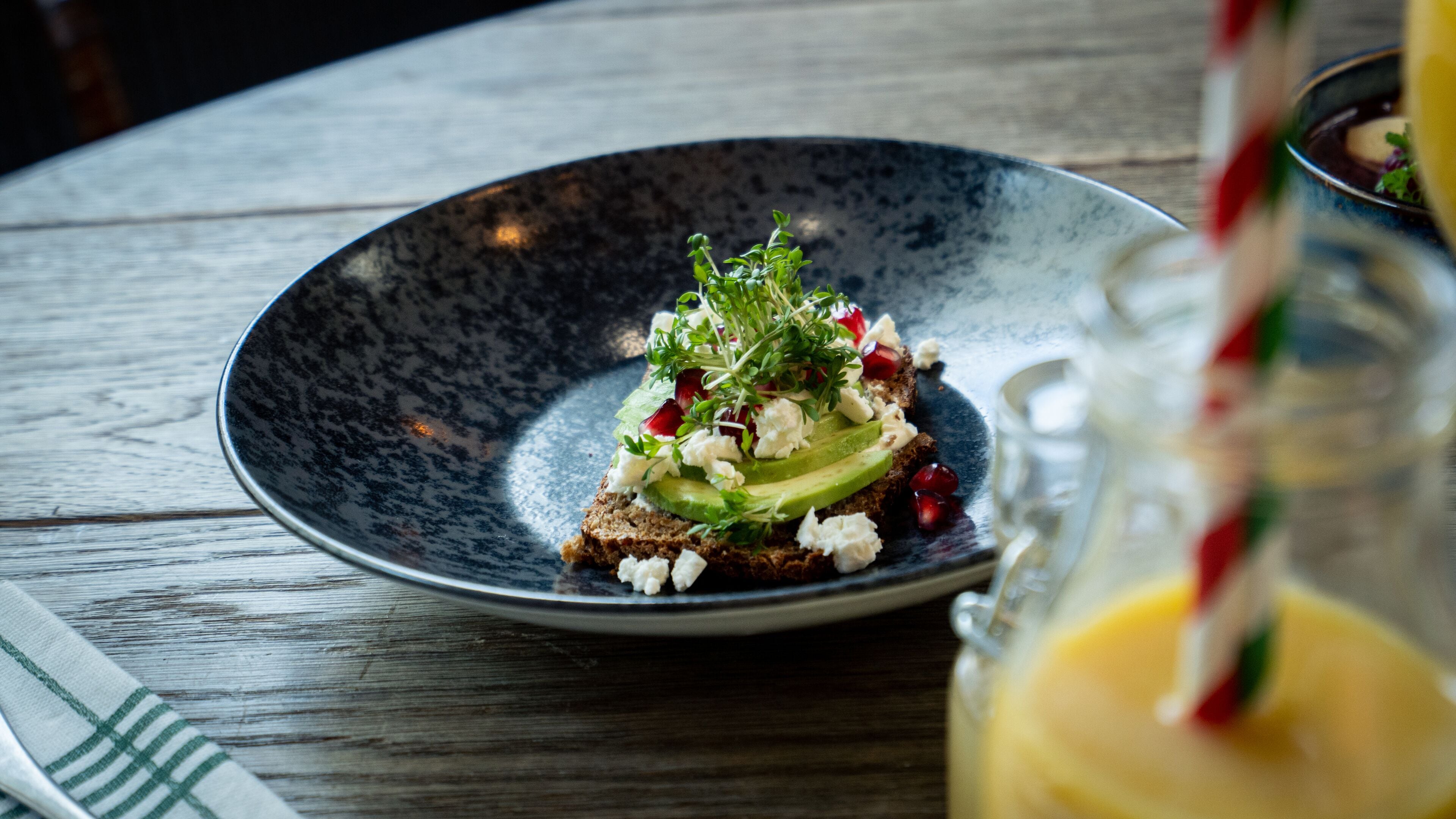 A dark ceramic plate with a slice of bread topped with avocado, feta cheese, and herbs, on a wooden table.