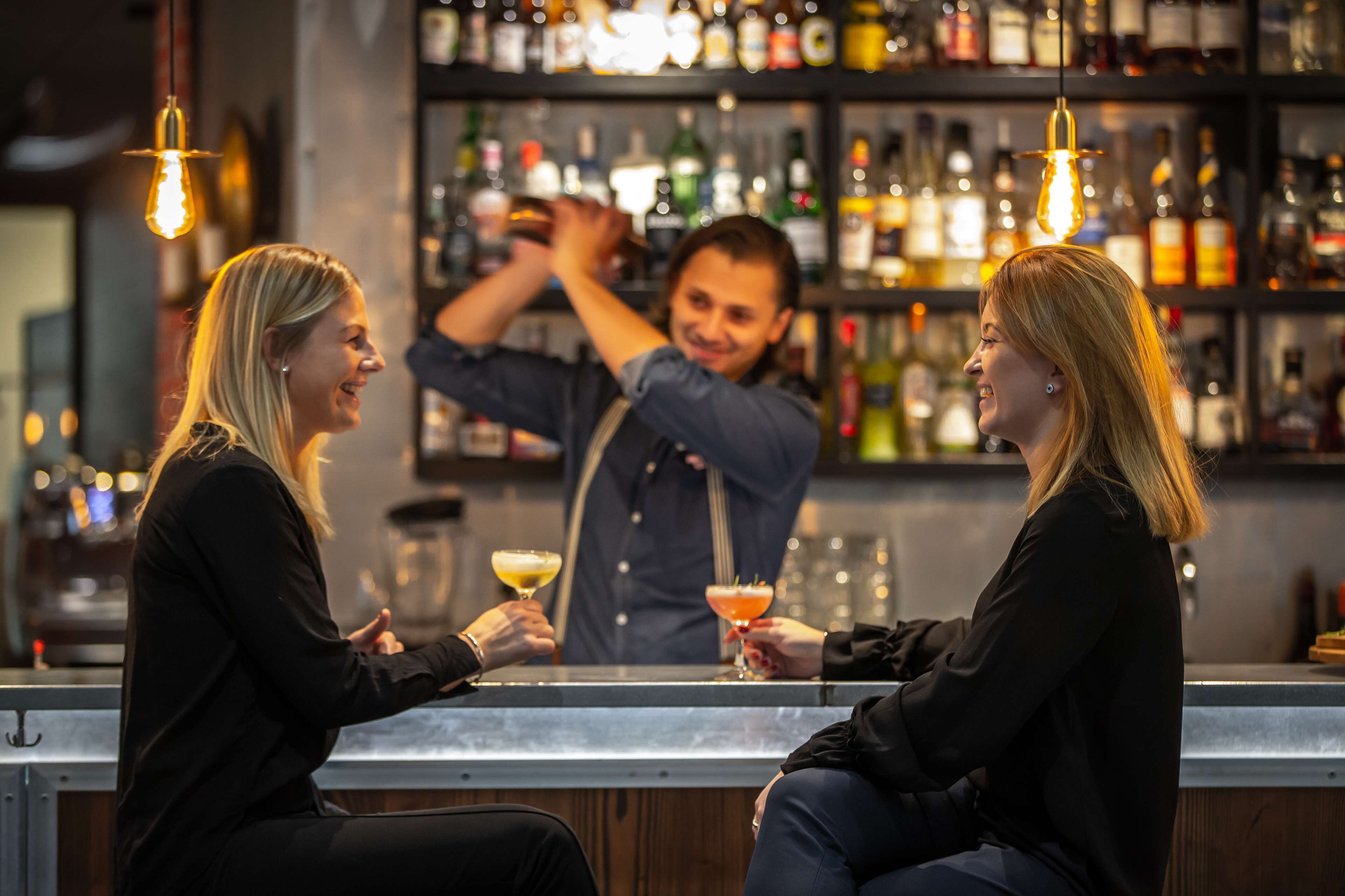 En bar med to personer siddende ved disken, en bartender der laver drinks, og hylder fyldt med flasker i baggrunden.