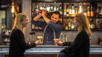 En bar med to personer siddende ved disken, en bartender der laver drinks, og hylder fyldt med flasker i baggrunden.