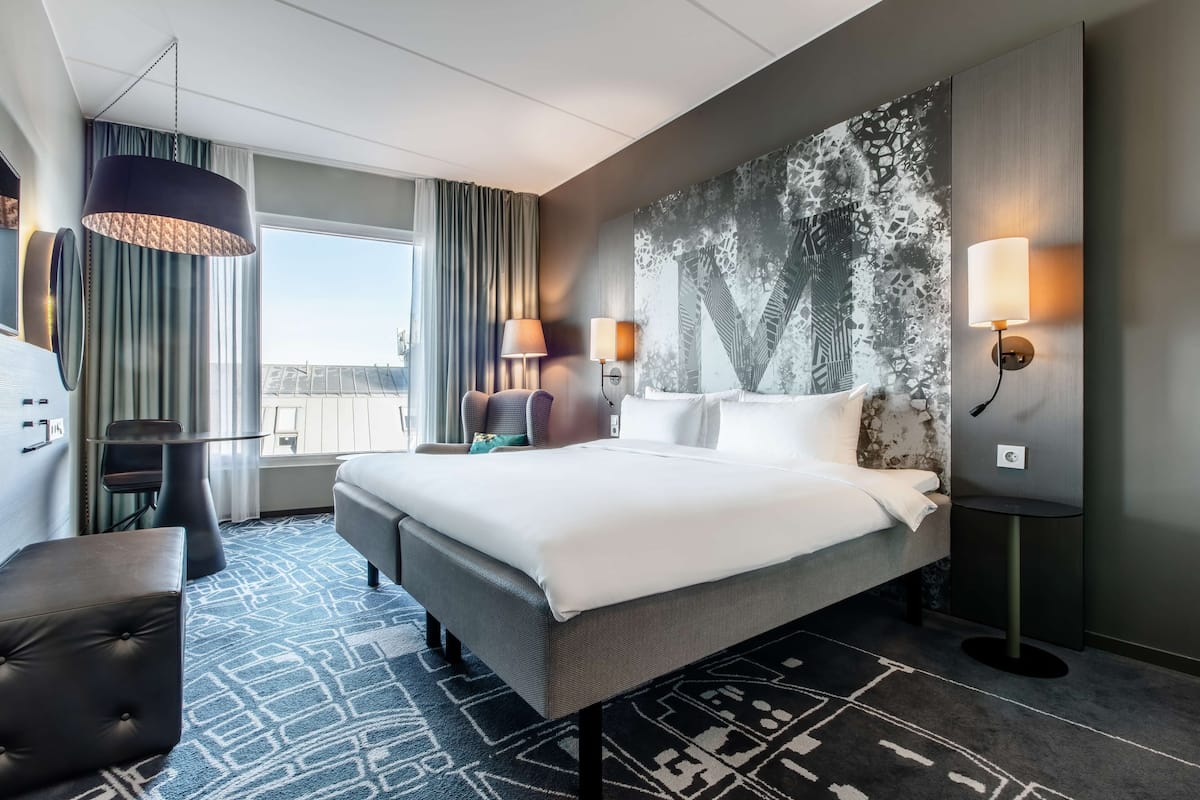 A hotel room with a large bed, two bedside tables, a desk, a chair, and a view of the city.