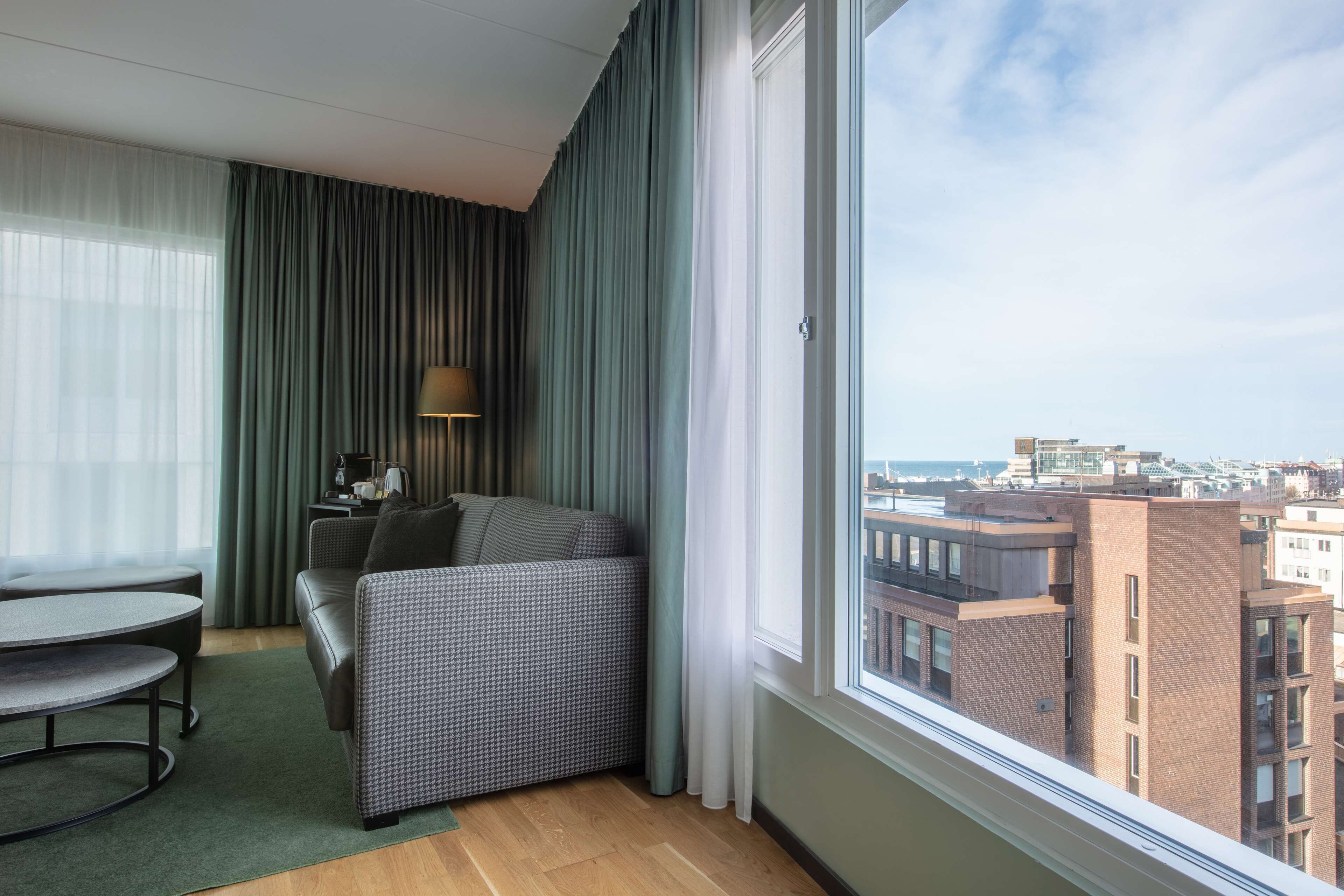 A modern hotel room with a view of the city, a sofa, a table, and a lamp.