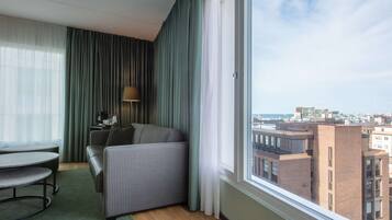 A modern hotel room with a view of the city, a sofa, a table, and a lamp.