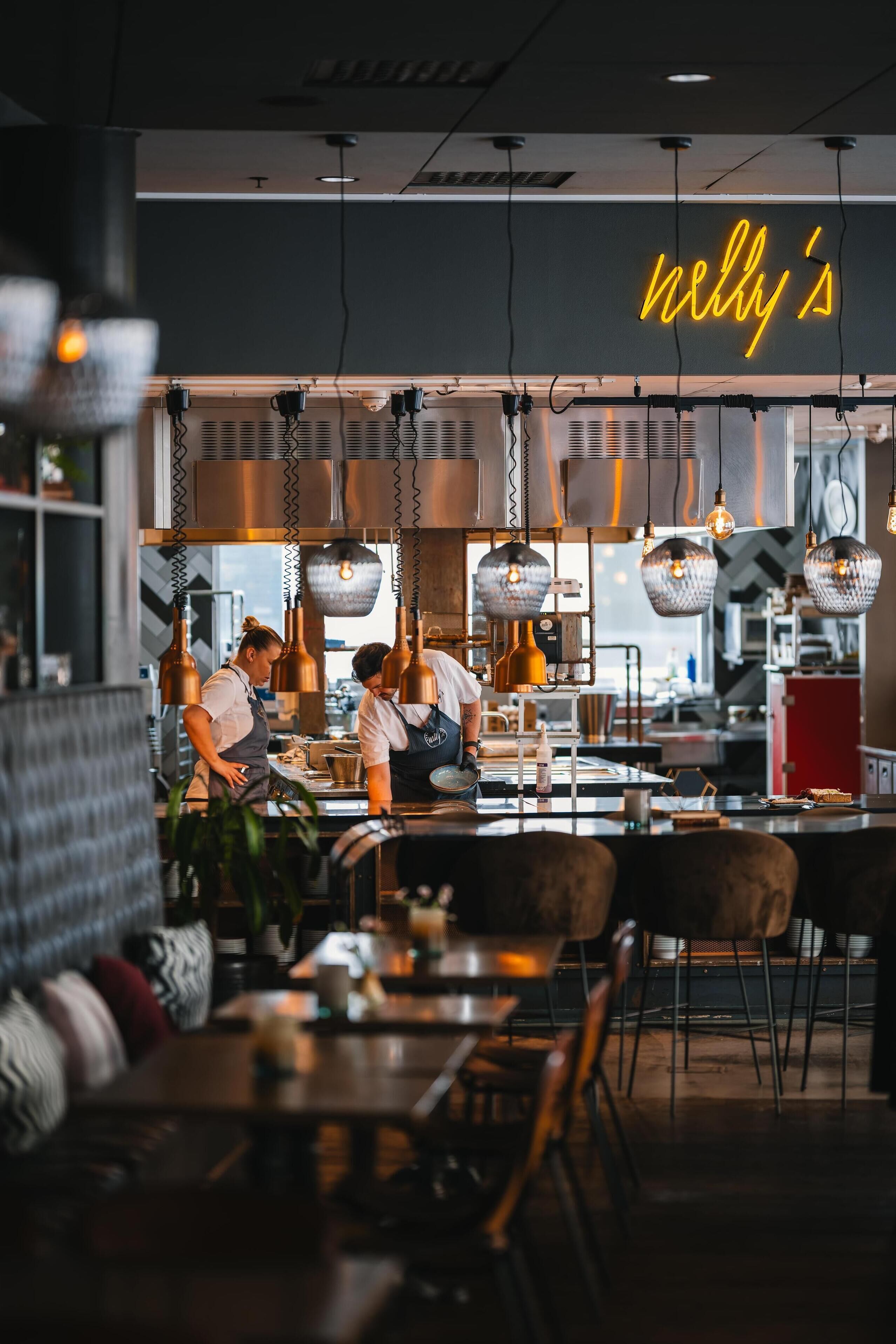 Een modern restaurantinterieur met hangende pendantlampen, een neonbord waarop 'nelly's' staat en een bar met krukken.