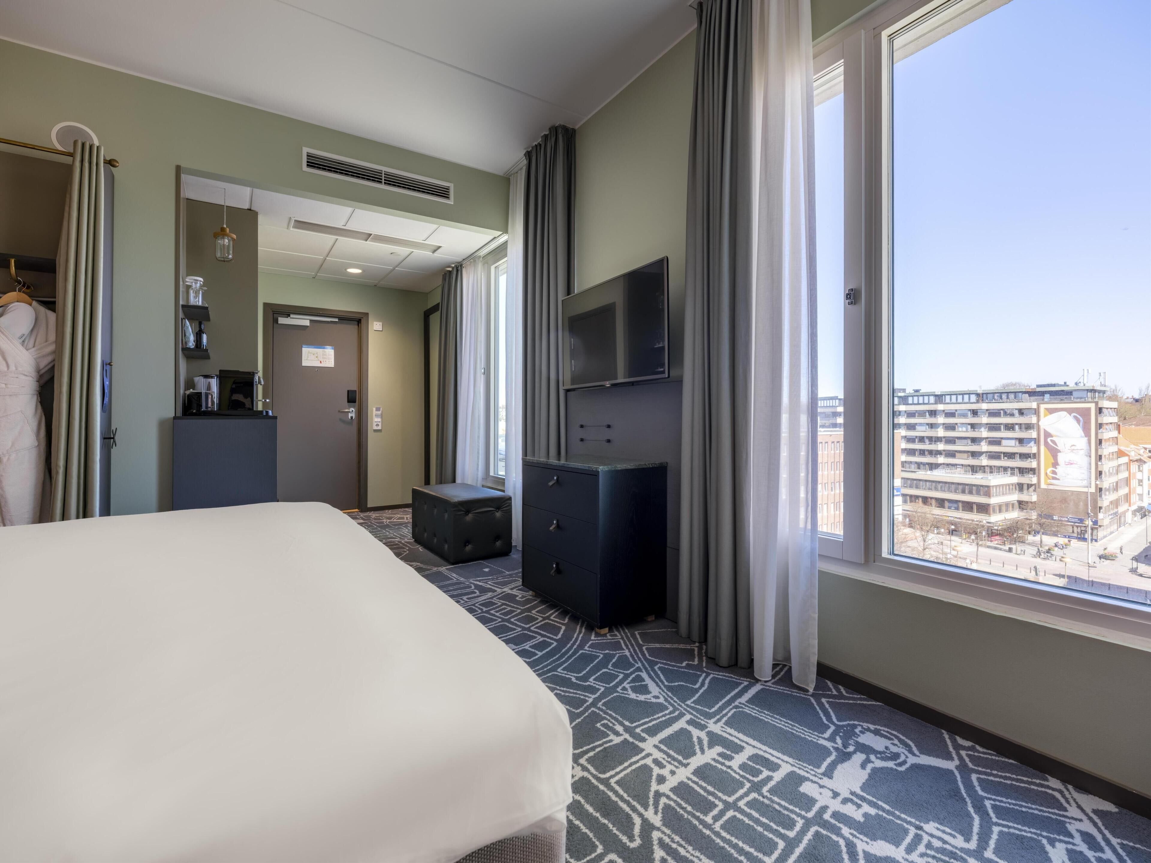 A hotel room with a bed, a television, a desk with a computer, a wardrobe, and a window with a city view.