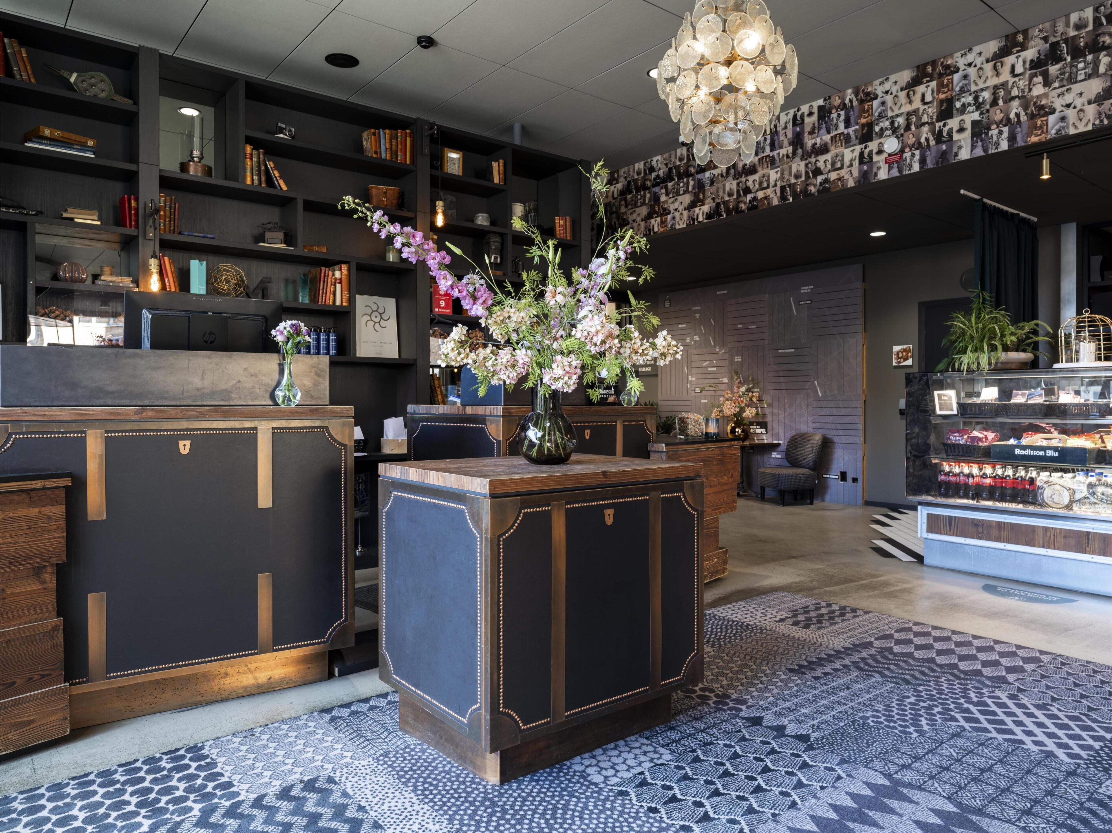 A reception desk with a vase of flowers, a bookshelf with books and decorative items, a wall of photographs, and a display case with snacks.