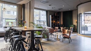 A modern hotel lobby with a long table, bar stools, a seating area with chairs, a bookshelf, and large windows offering a view of the street. - Radisson Blu Metropol Hotel, Helsingborg (Helsingborg)