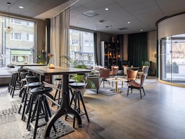 Un moderno vestíbulo de hotel con una mesa larga, taburetes de bar, una zona de estar con sillas, una estantería y grandes ventanales con vistas a la calle.