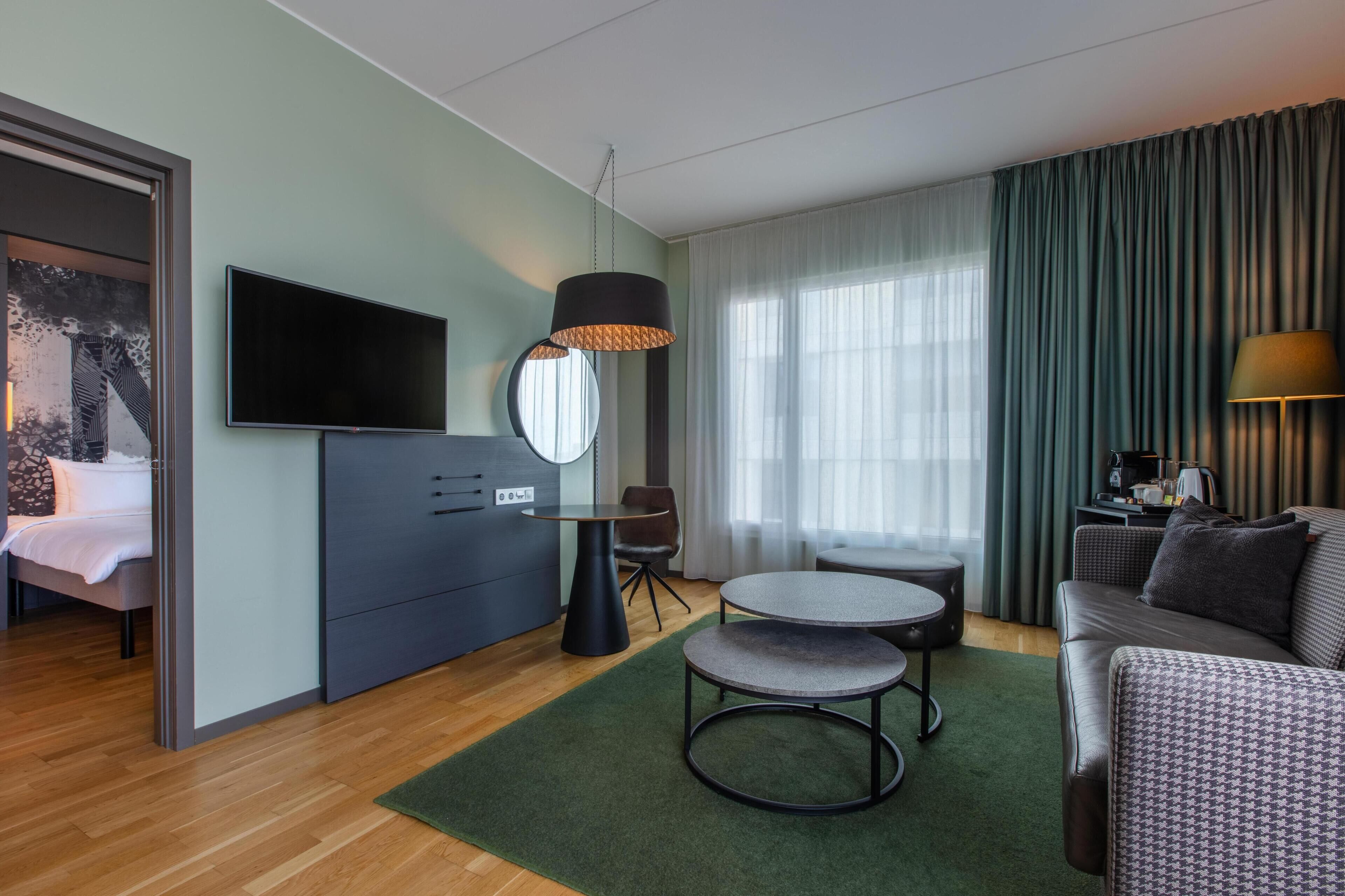 A modern hotel room with a bed, a TV, a desk, a round table, a sofa, and a large window with curtains.