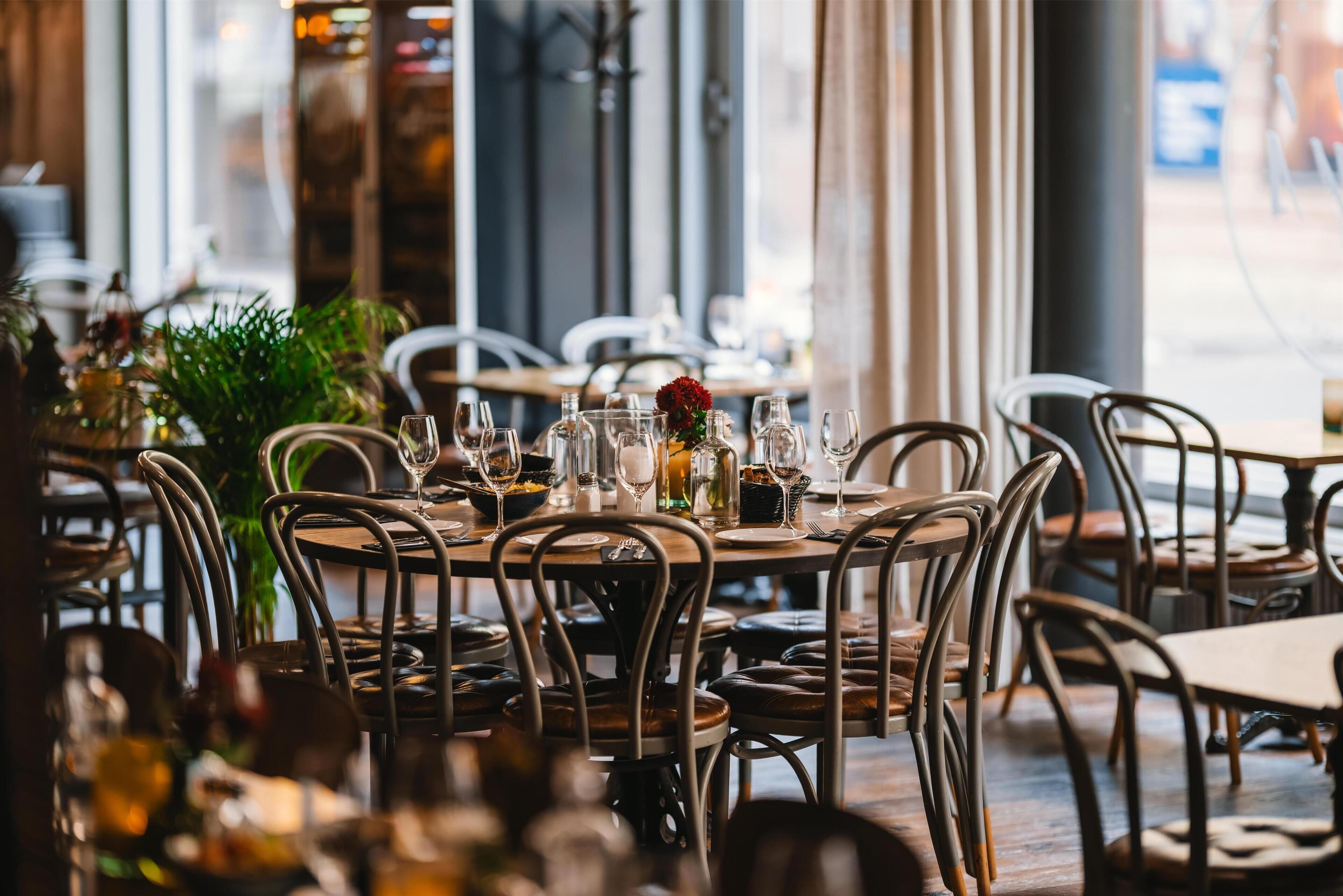 Ein Restaurantinneres mit runden Tischen, Metallstühlen und einem großen Fenster.