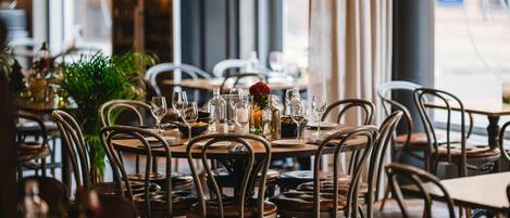 Ein Restaurantinneres mit runden Tischen, Metallstühlen und einem großen Fenster.