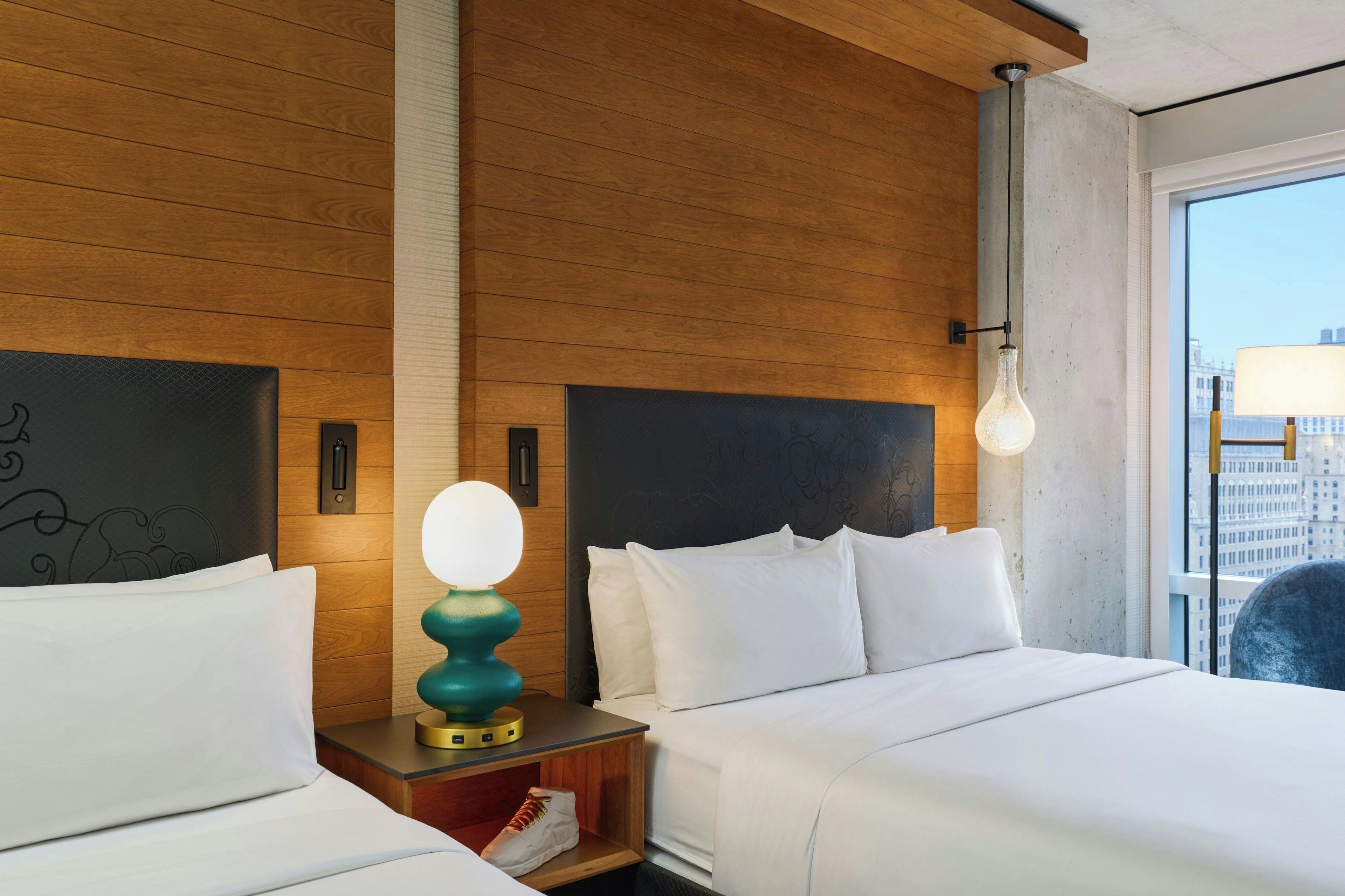 Ein Hotelzimmer mit Holzwand, einem Bett mit weißer Bettwäsche, einem Nachttisch mit Lampe und Blick auf die Stadt durch ein großes Fenster.
