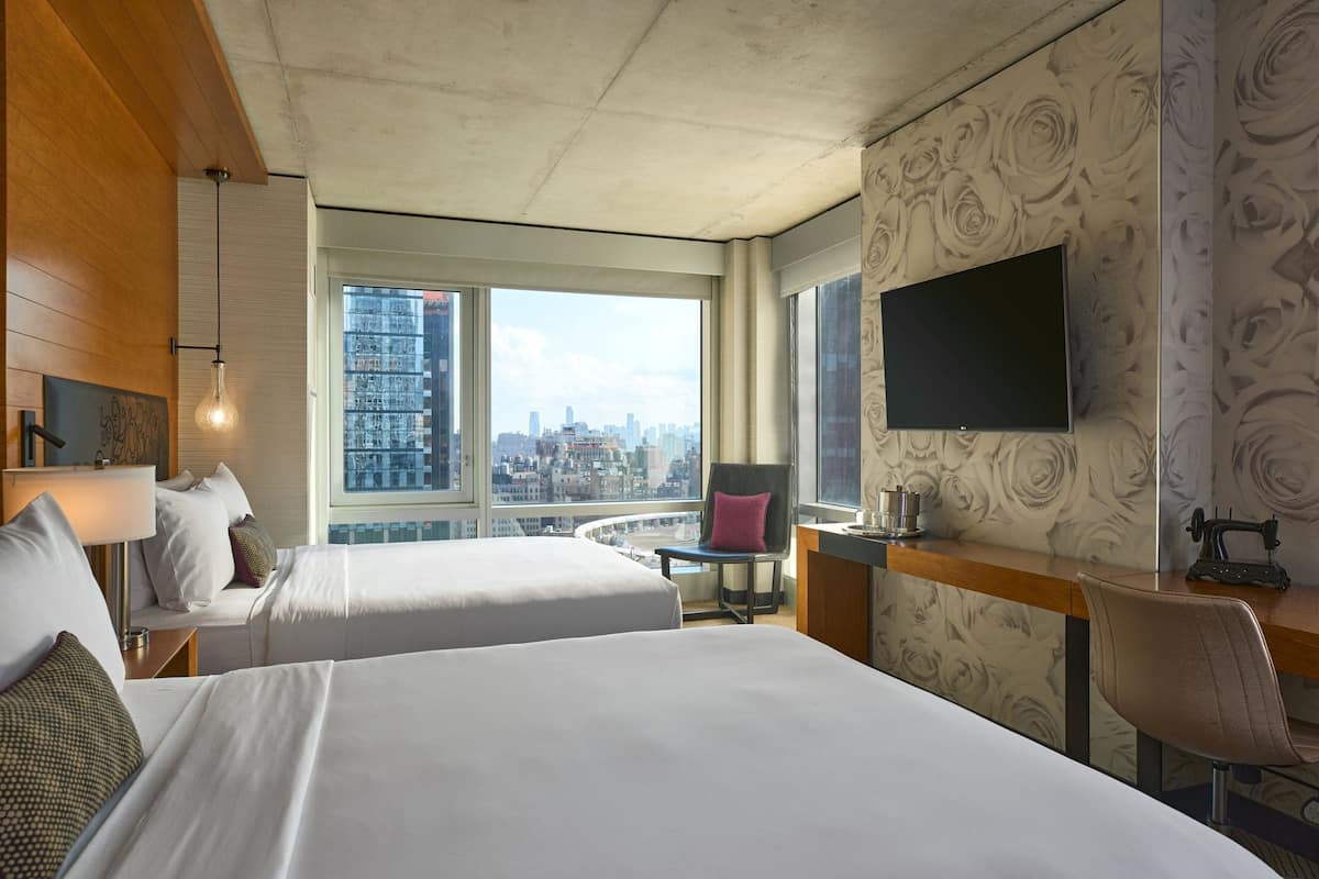 Habitación de hotel con dos camas, un escritorio, una silla, un televisor y un ventanal con vistas a la ciudad.