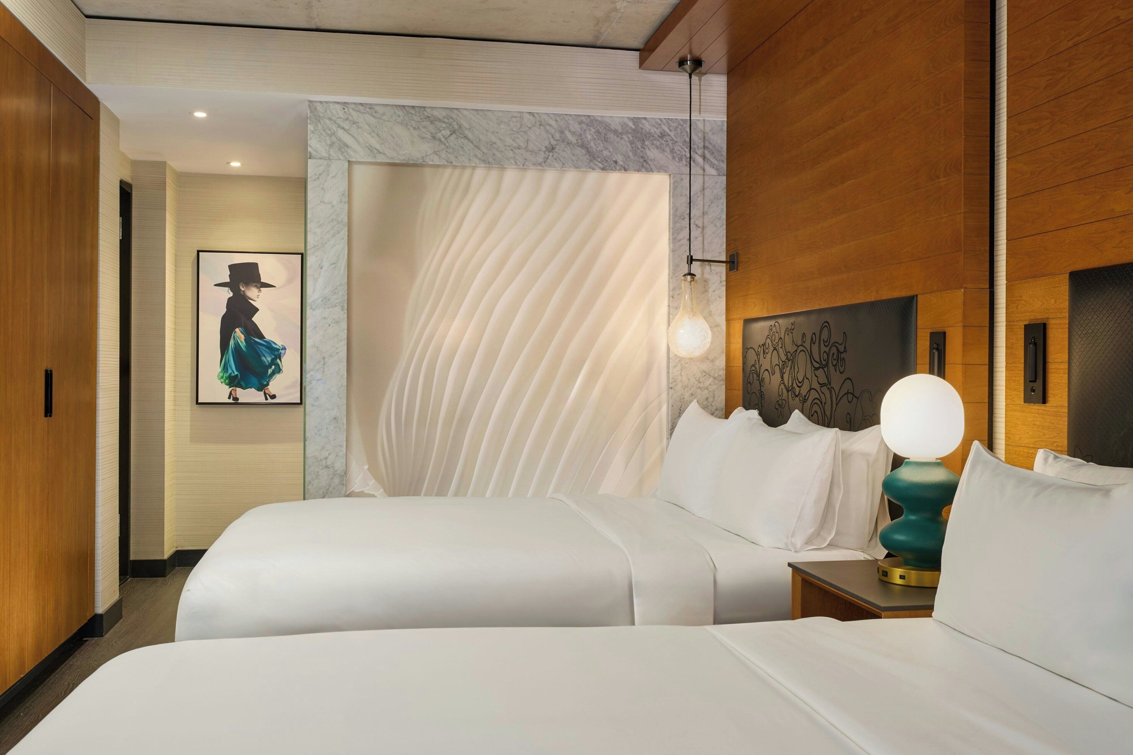 A hotel room with two beds, a marble wall, a framed artwork, and a modern light fixture.