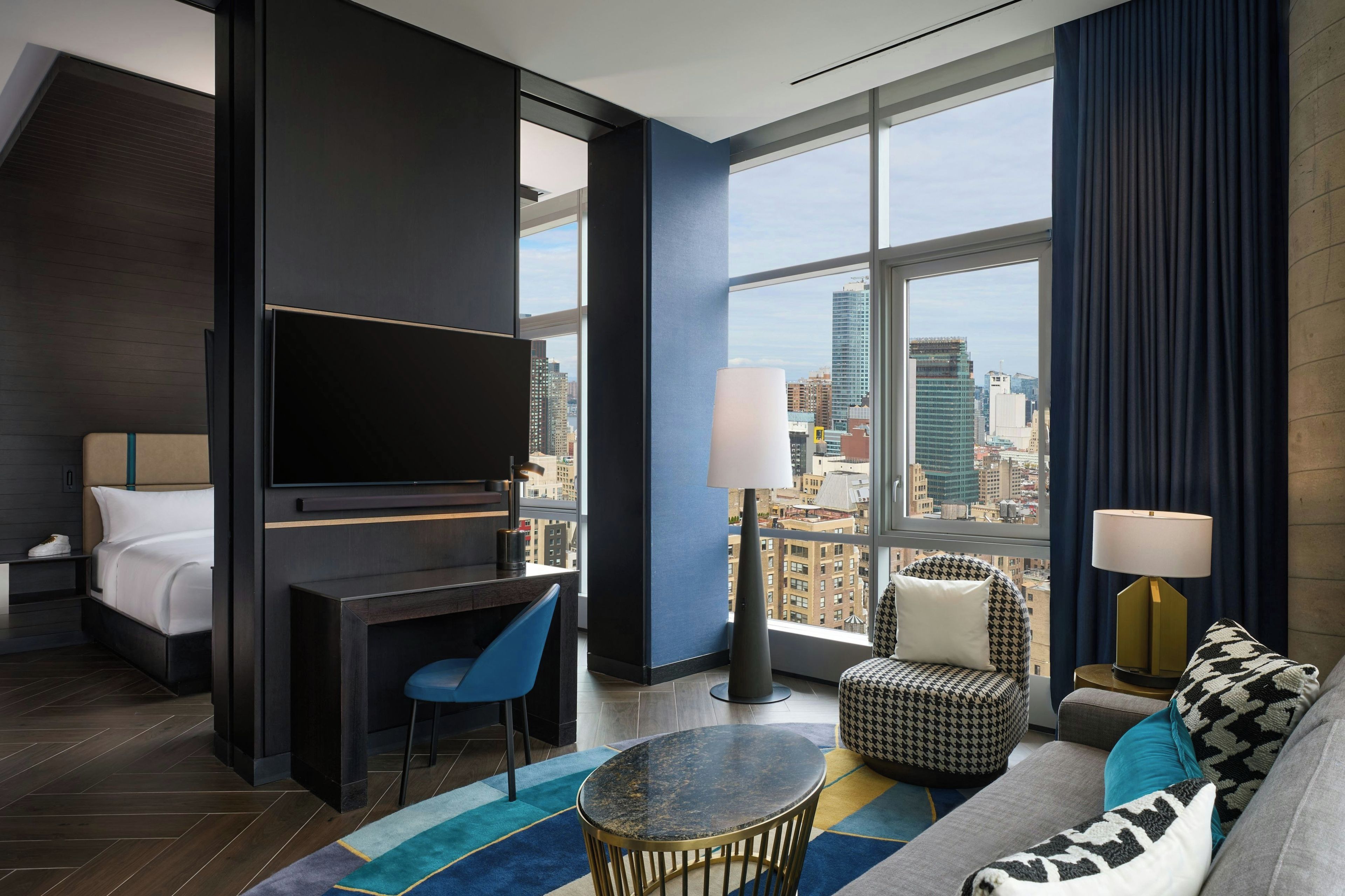 A modern hotel room with a bed, a desk, a TV, a sofa, and a view of the cityscape.