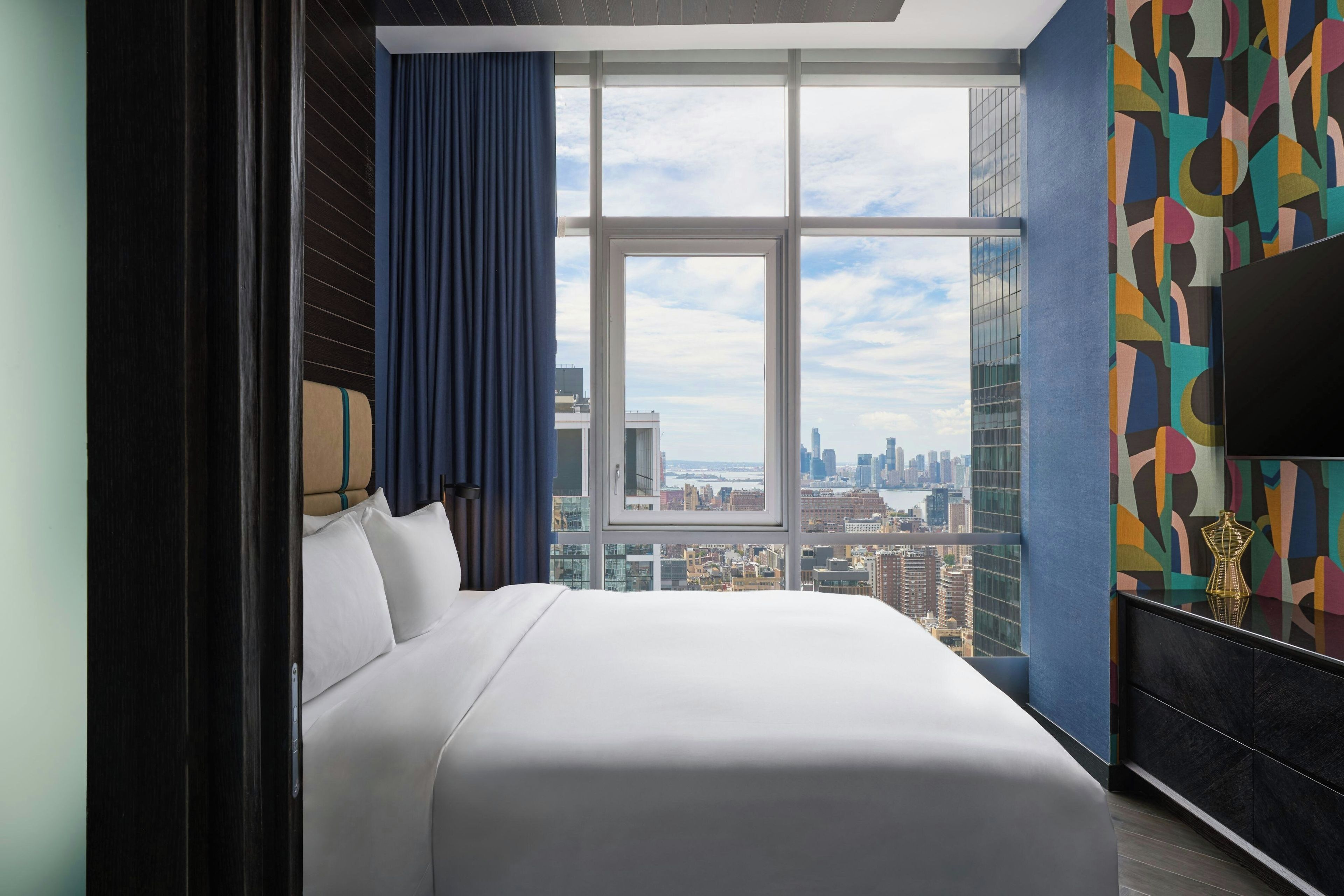 Camera d'albergo con un letto grande, vista sulla città attraverso una finestra a tutta altezza e una parete moderna con motivi geometrici.