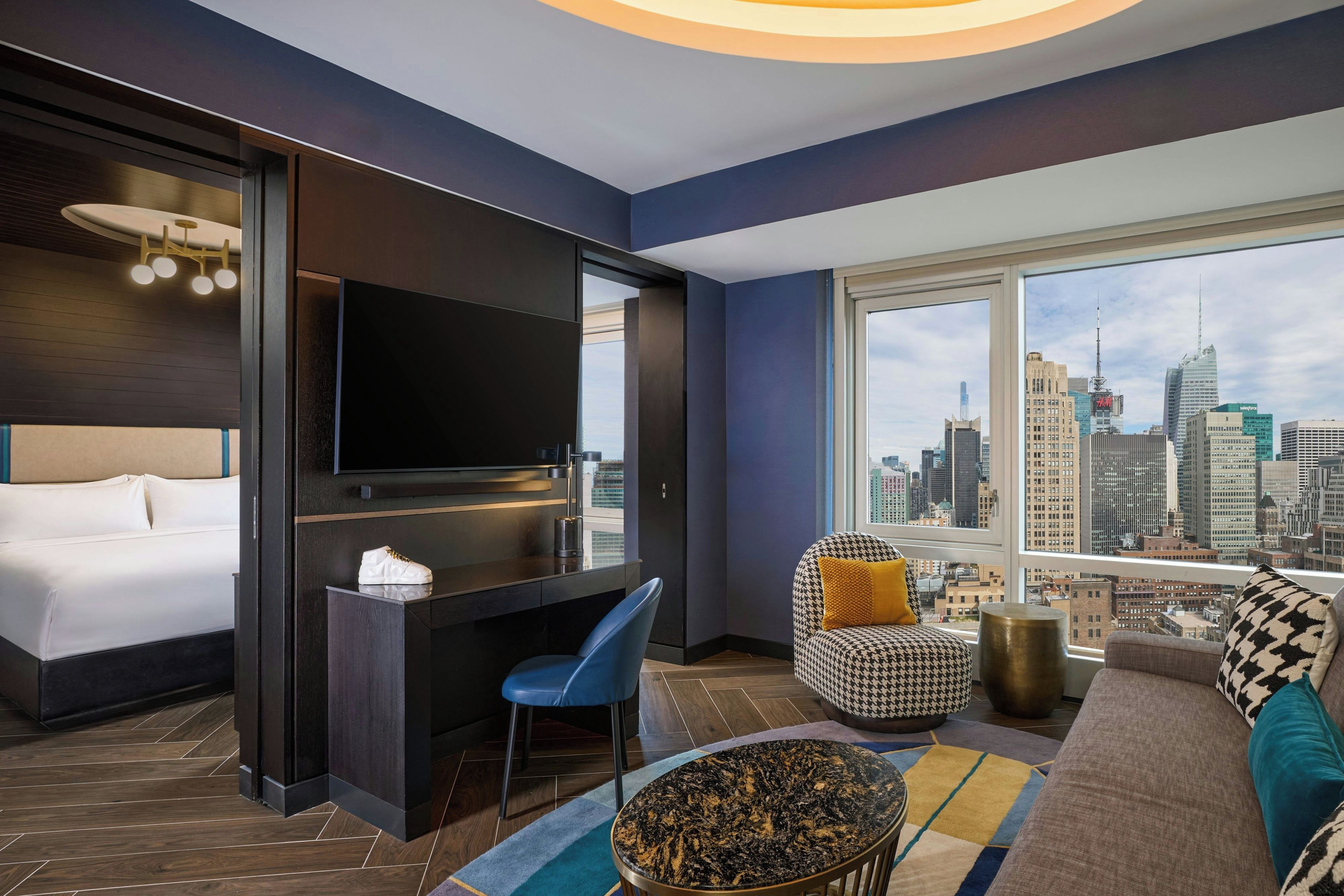 Habitación de hotel moderna con cama, escritorio, silla, sofá, televisor y una ventana grande con vistas a la ciudad.