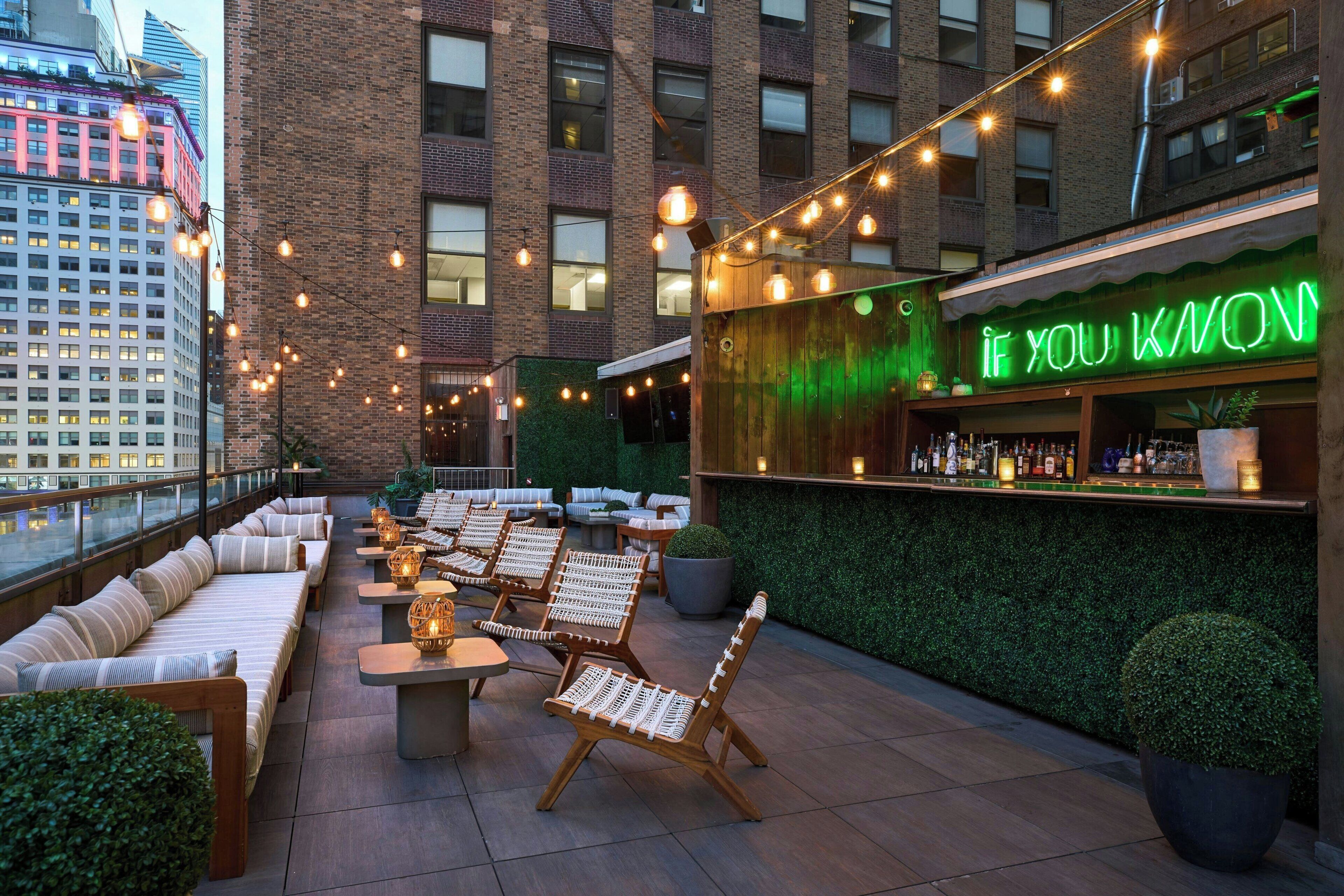 Uma área externa com assentos acolchoados, mesas, um bar com letreiro de neon e uma parede verde com plantas.