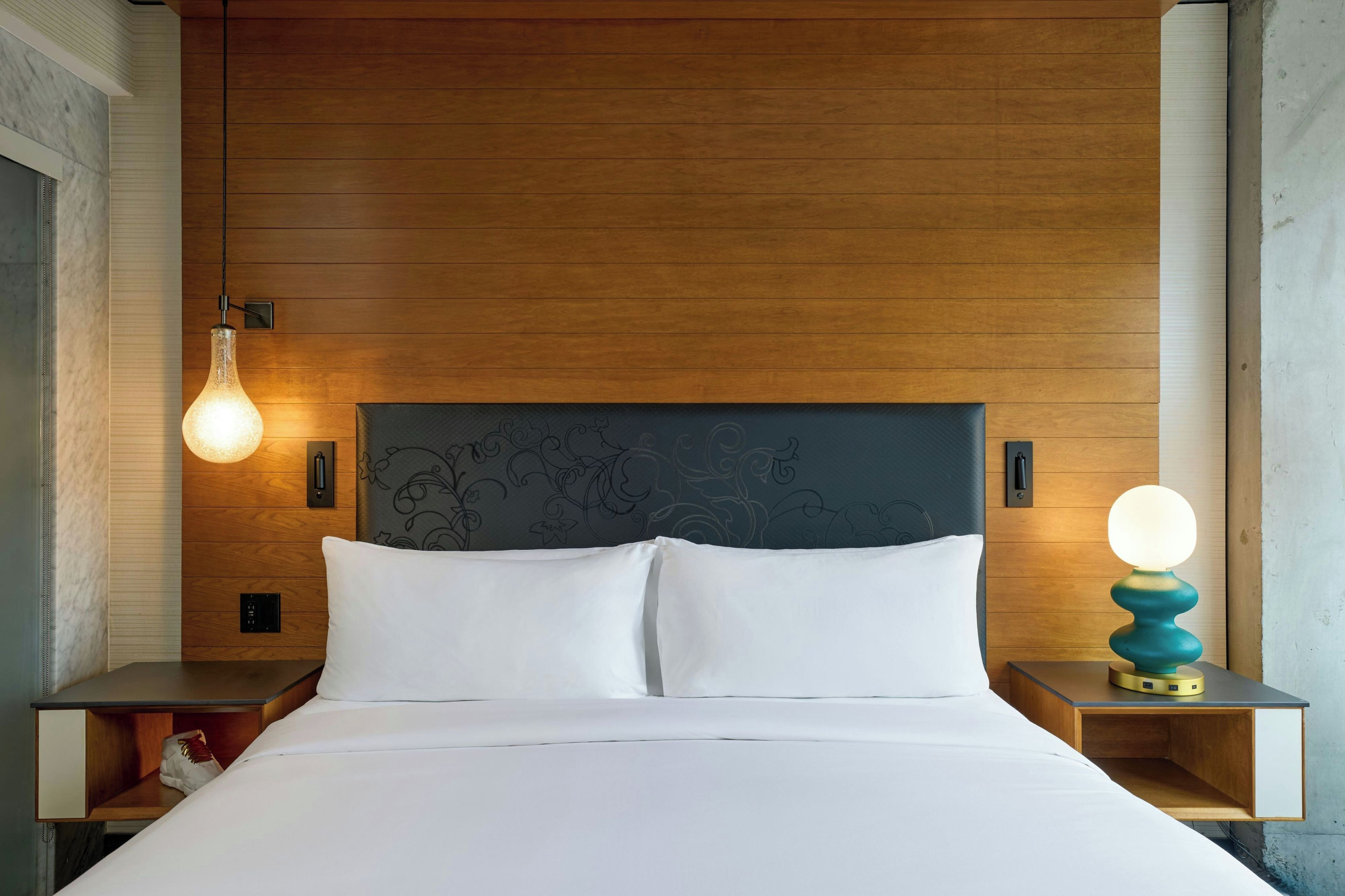 A hotel room with a wooden headboard, a bed with white linens, a nightstand with a lamp, and a bedside table with a decorative lamp.