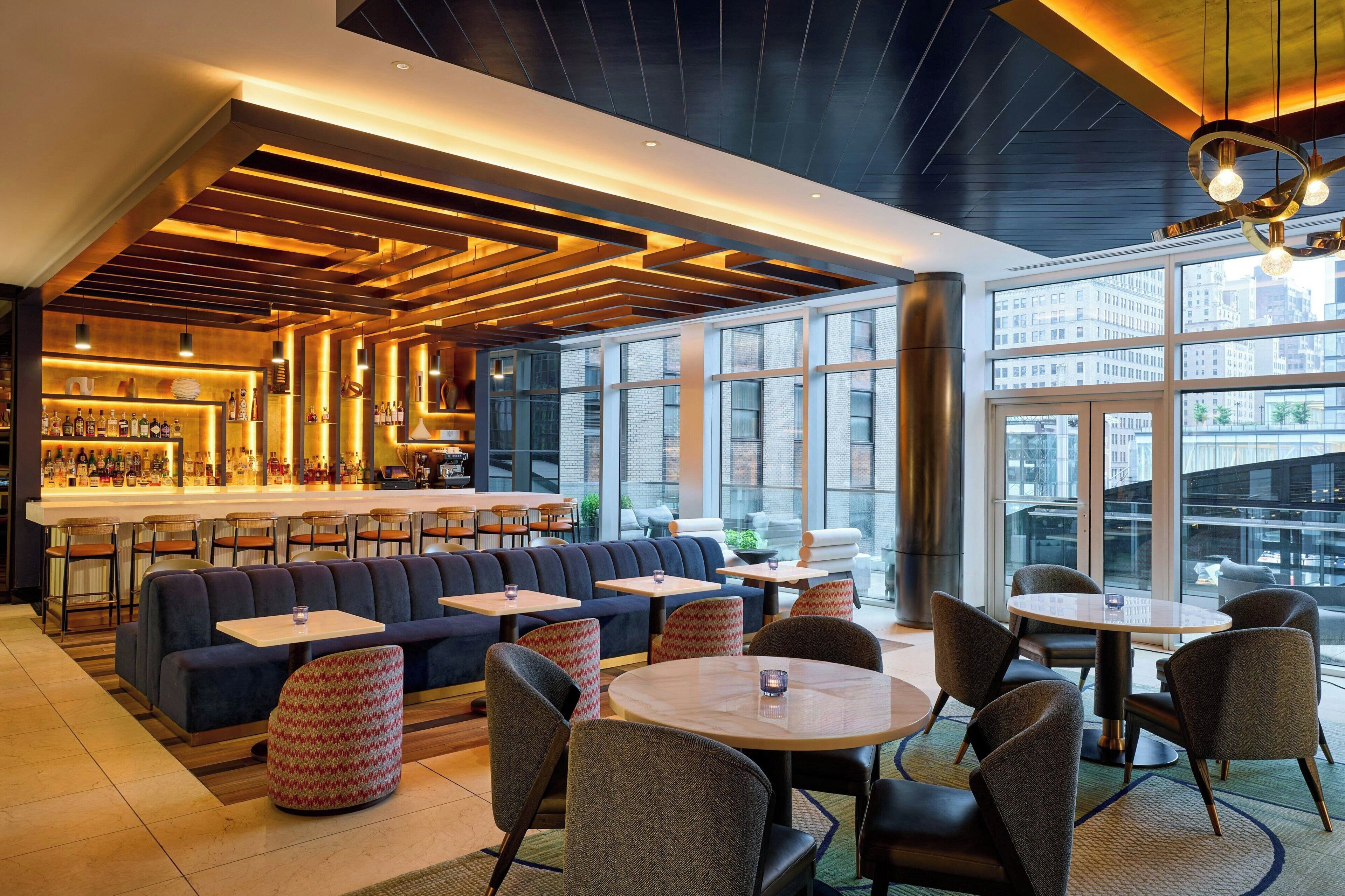 A modern bar area with seating, a bar counter, and large windows offering city views.