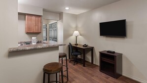 A compact kitchen with a countertop, a coffee maker, a chair, a desk with a lamp, and a TV mounted on the wall. - Eagle's Den Suites Big Spring a Travelodge by Wyndham (Big Spring)