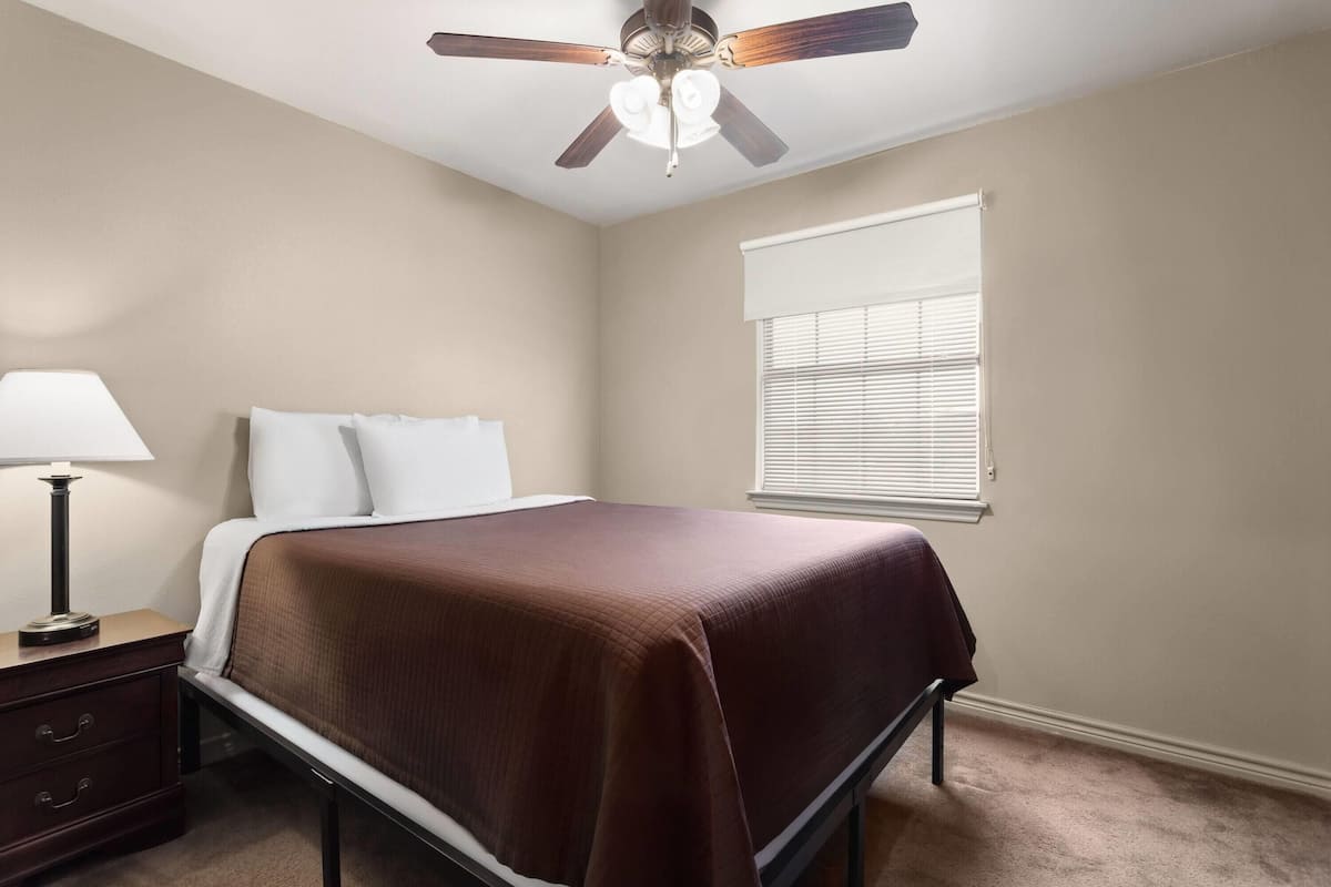Un dormitorio con cama, mesita de noche, ventilador de techo y ventana con persianas.