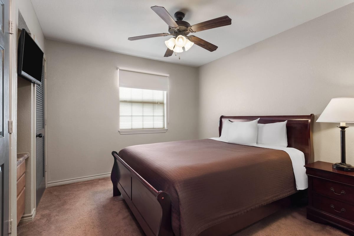 Un dormitorio con cama, ventilador de techo, mesita de noche, lámpara, ventana y puerta.