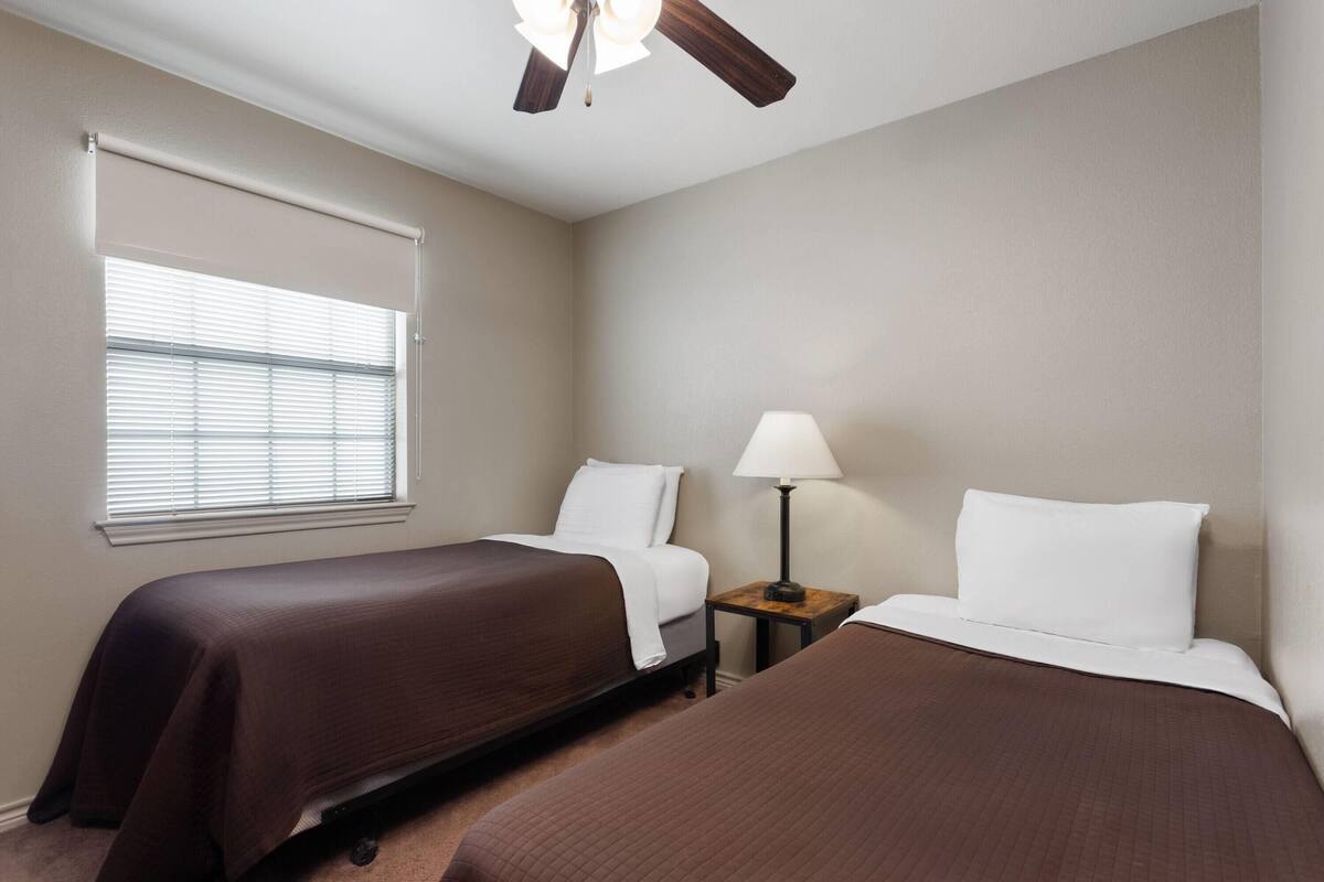 Habitación de hotel con dos camas, un ventilador de techo, una ventana con persianas y una mesita de noche con lámpara.